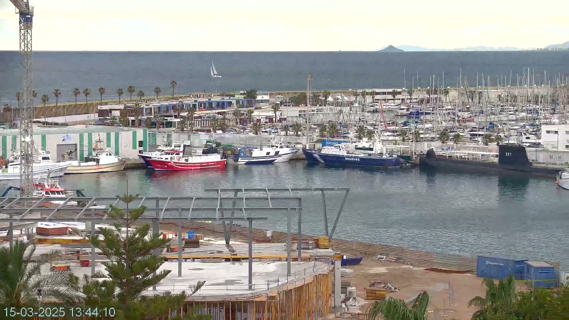 A harbor filled with numerous boats, including fishing vessels and a submarine, is partially visible under partly cloudy skies, with construction underway nearby.