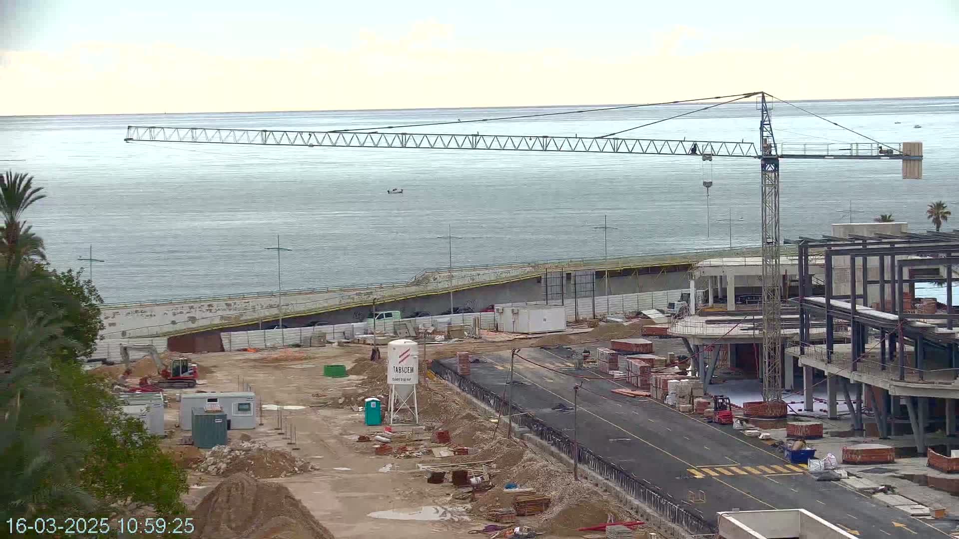 A construction site near a body of water under an overcast sky features a large crane, several buildings under construction, and various construction materials scattered around.