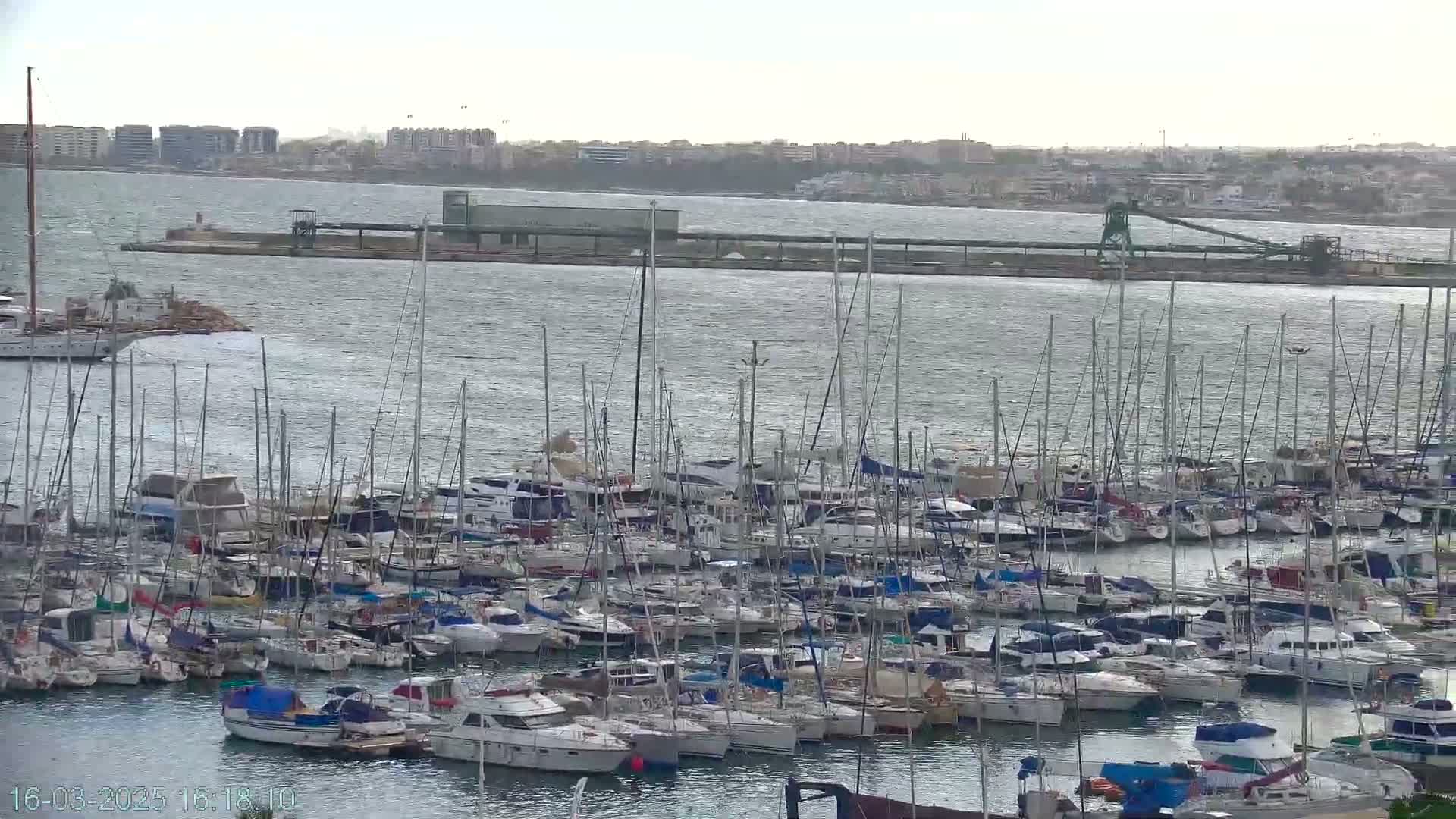 A large marina filled with numerous sailboats and motorboats is visible, situated on a body of water under somewhat overcast skies.