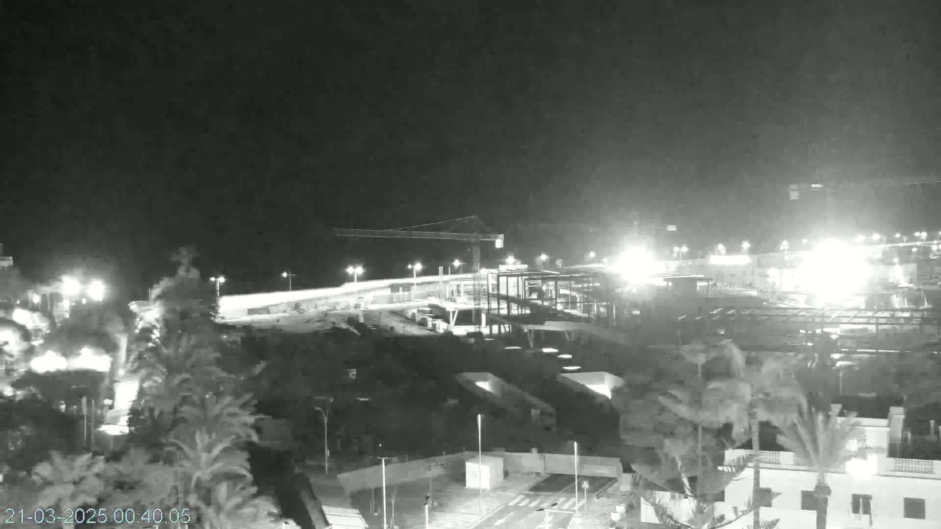 A nighttime, black and white view of a construction site near a coast, illuminated by artificial lights under a dark sky.