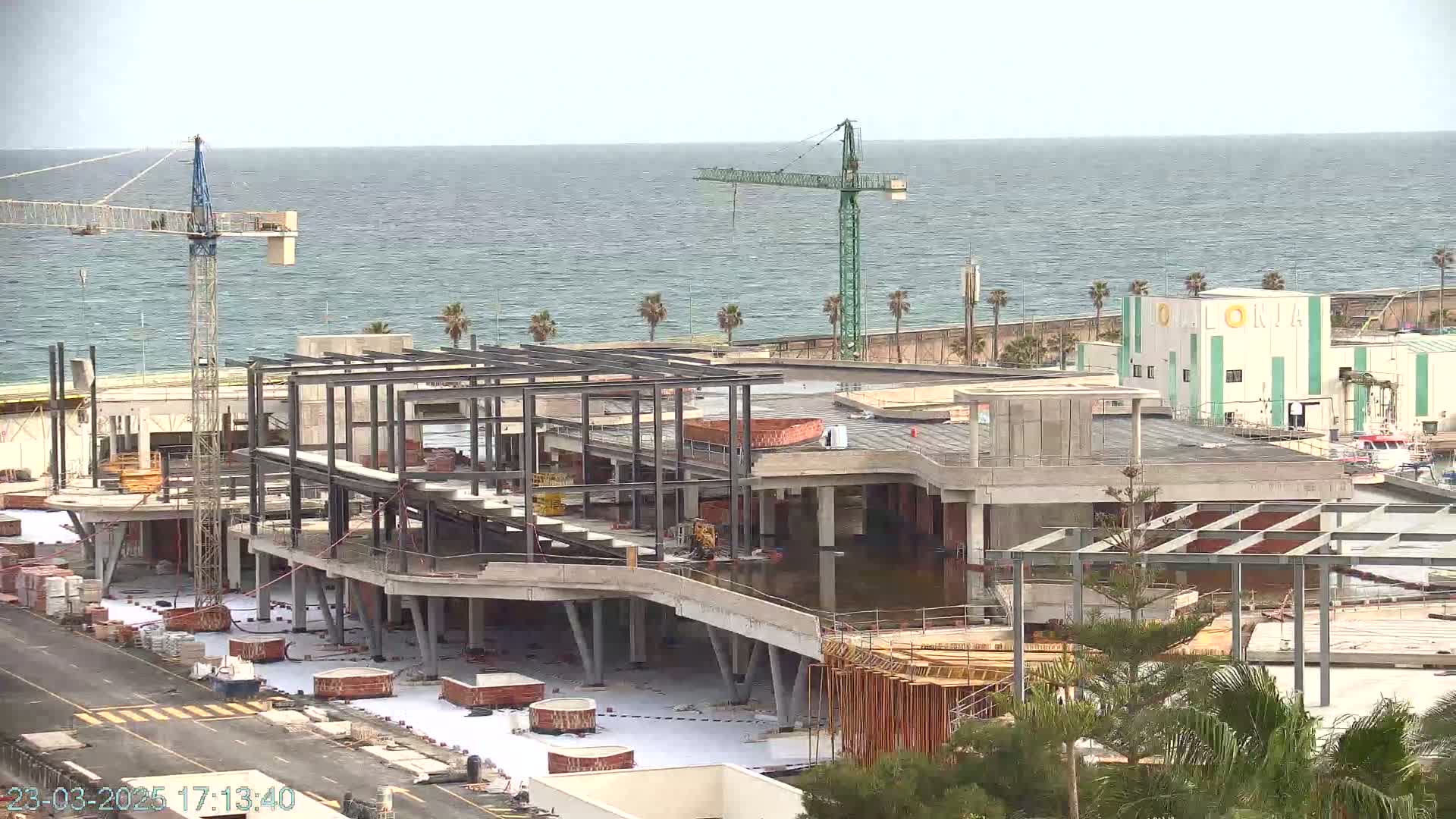 A large building under construction near a body of water on an overcast day, with two cranes visible on the site.