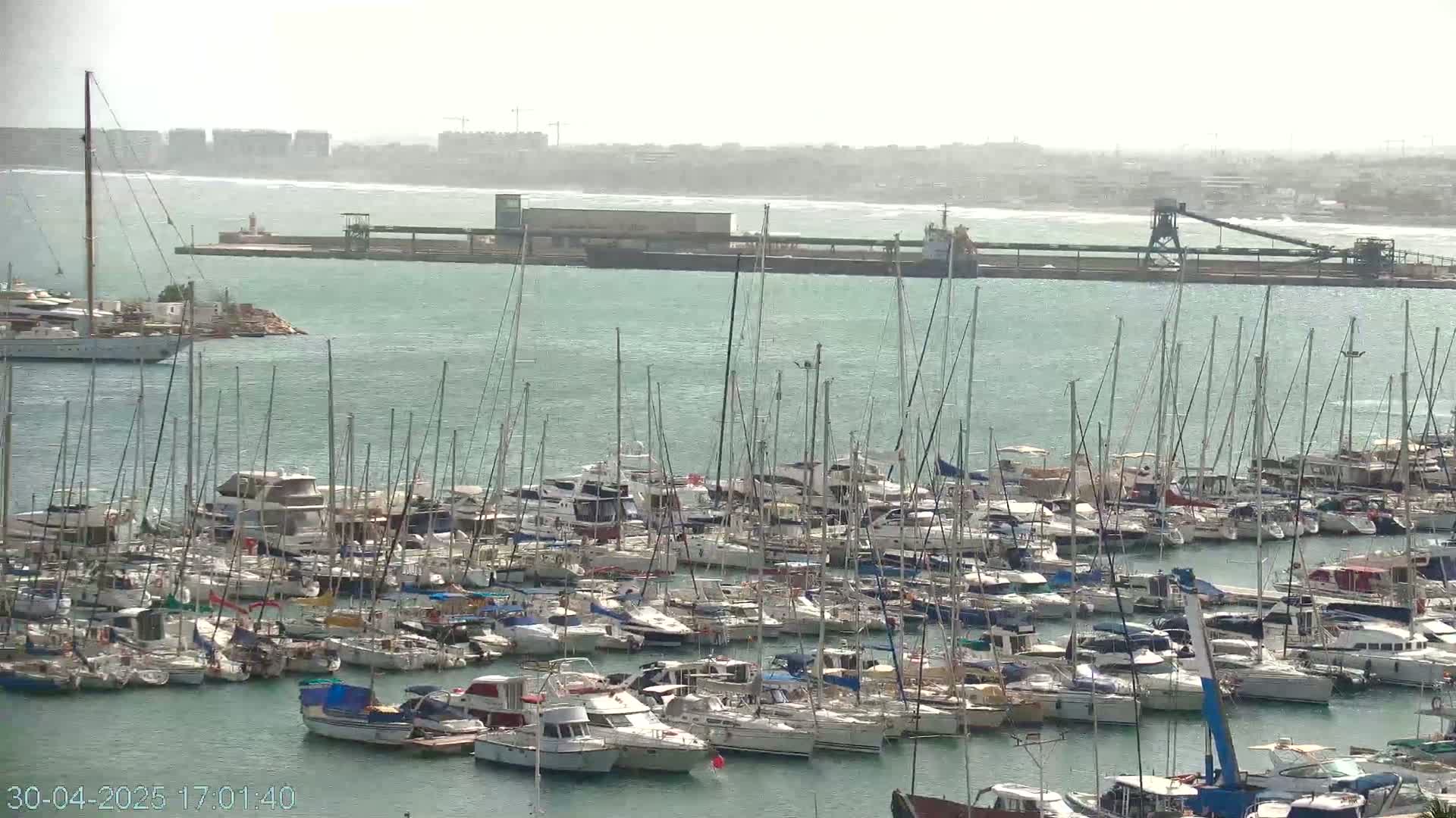A hazy, sunny day reveals a marina filled with numerous sailboats and motorboats, with a larger vessel and industrial structures visible in the distance across the water.