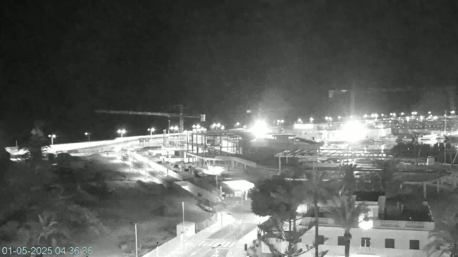 A nighttime, black and white view of a coastal construction site with bright lights illuminating the area, next to a dark body of water.
