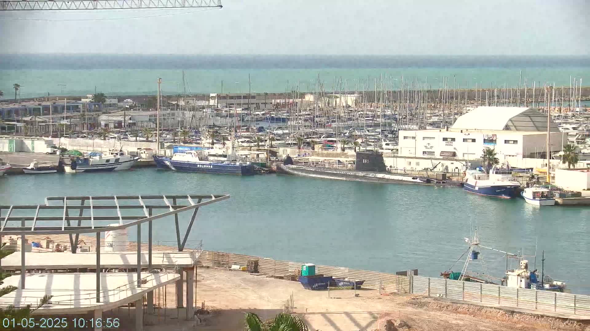 A marina filled with numerous boats and a submarine, under a mostly clear sky with a calm sea in the background.