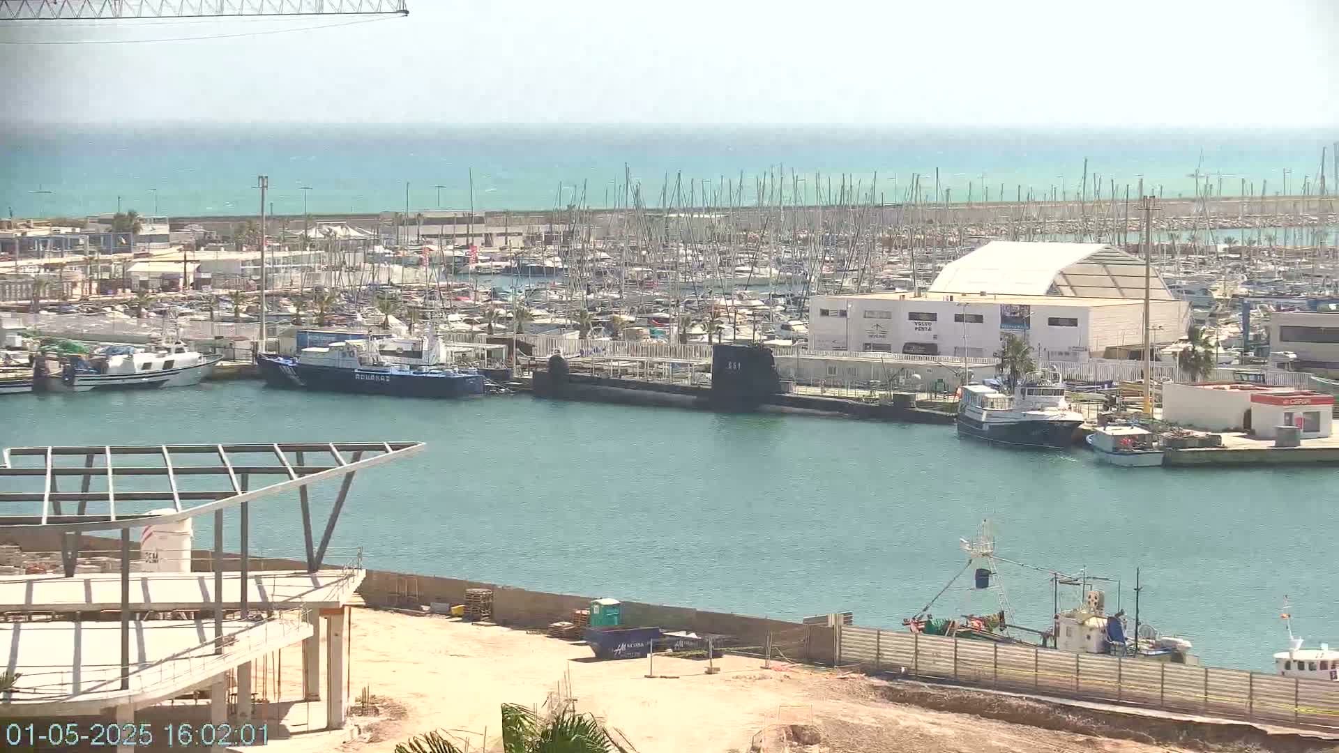A harbor filled with numerous boats and a submarine, under a clear, sunny sky, with construction visible in the foreground.