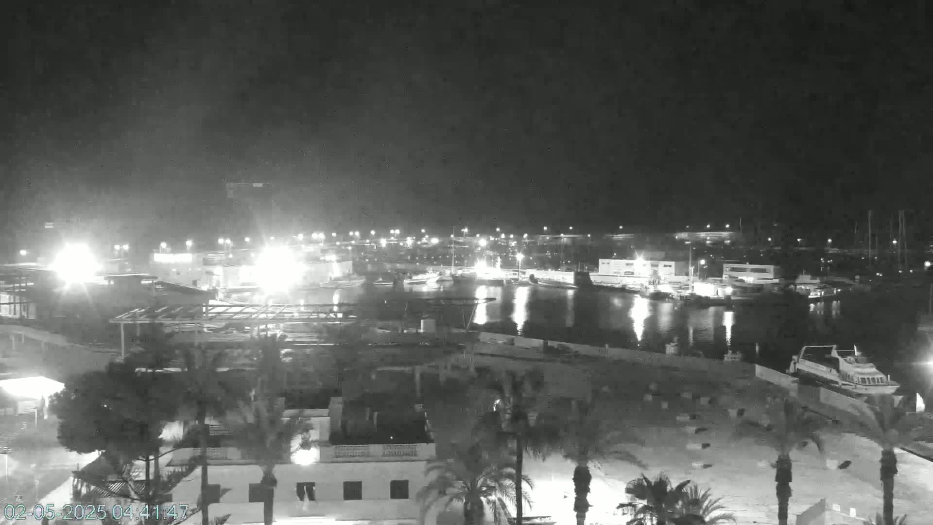 A nighttime, high-angle view of a marina with boats docked, surrounded by buildings and palm trees under a dark sky.