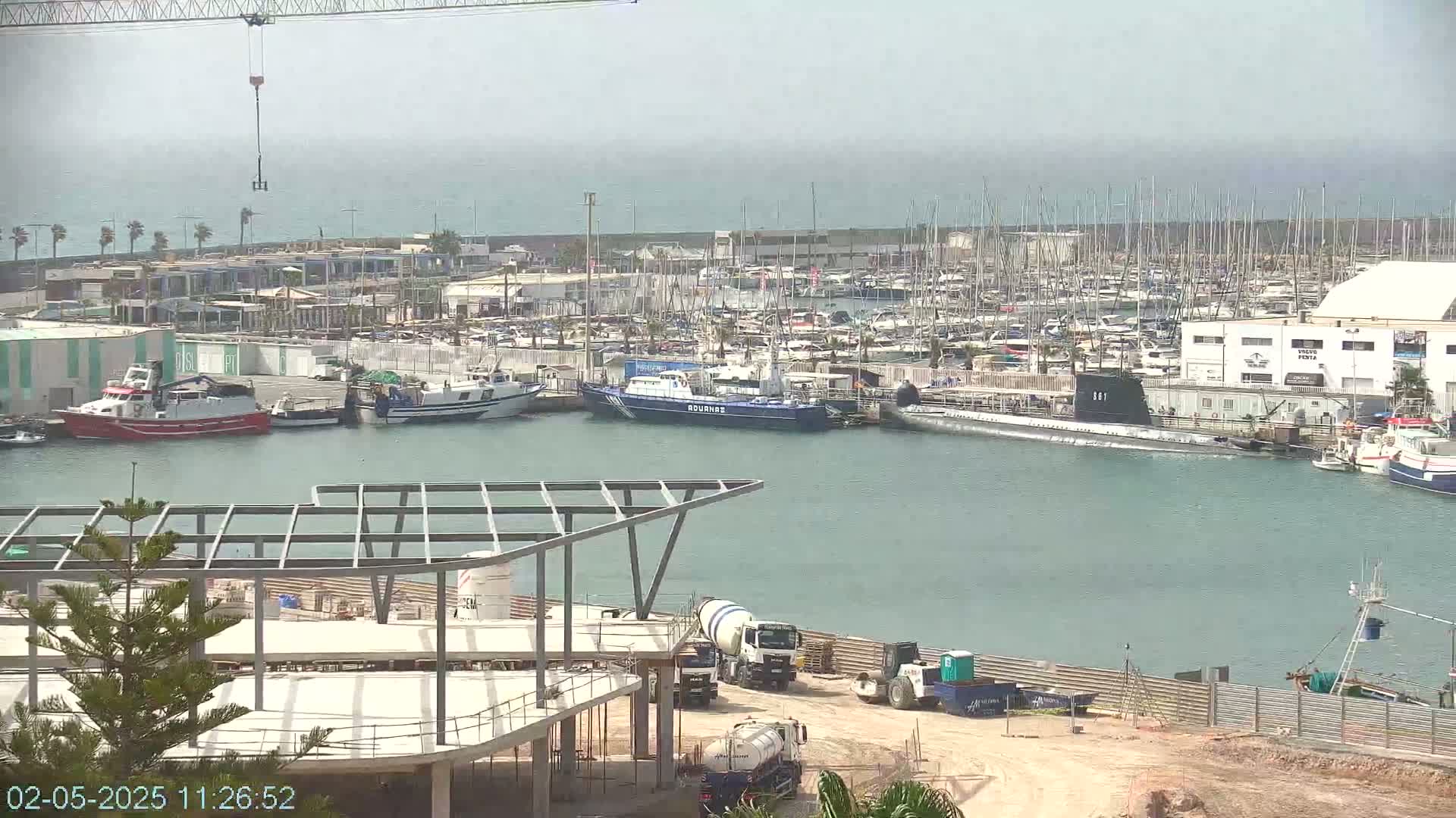 A harbor filled with numerous boats and a docked submarine, under a hazy sky, is viewed from above a construction site.