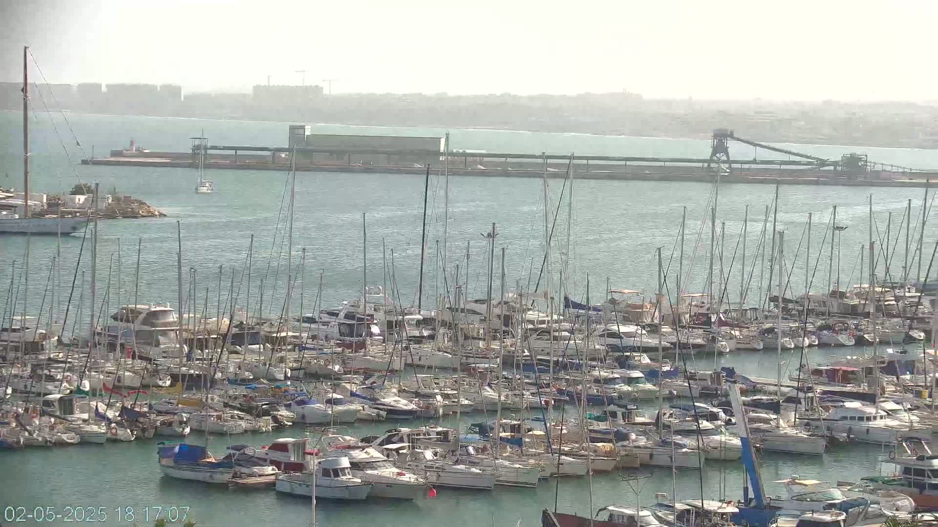 A hazy harbor filled with numerous sailboats and motorboats docked closely together, with a distant industrial area visible across the water.