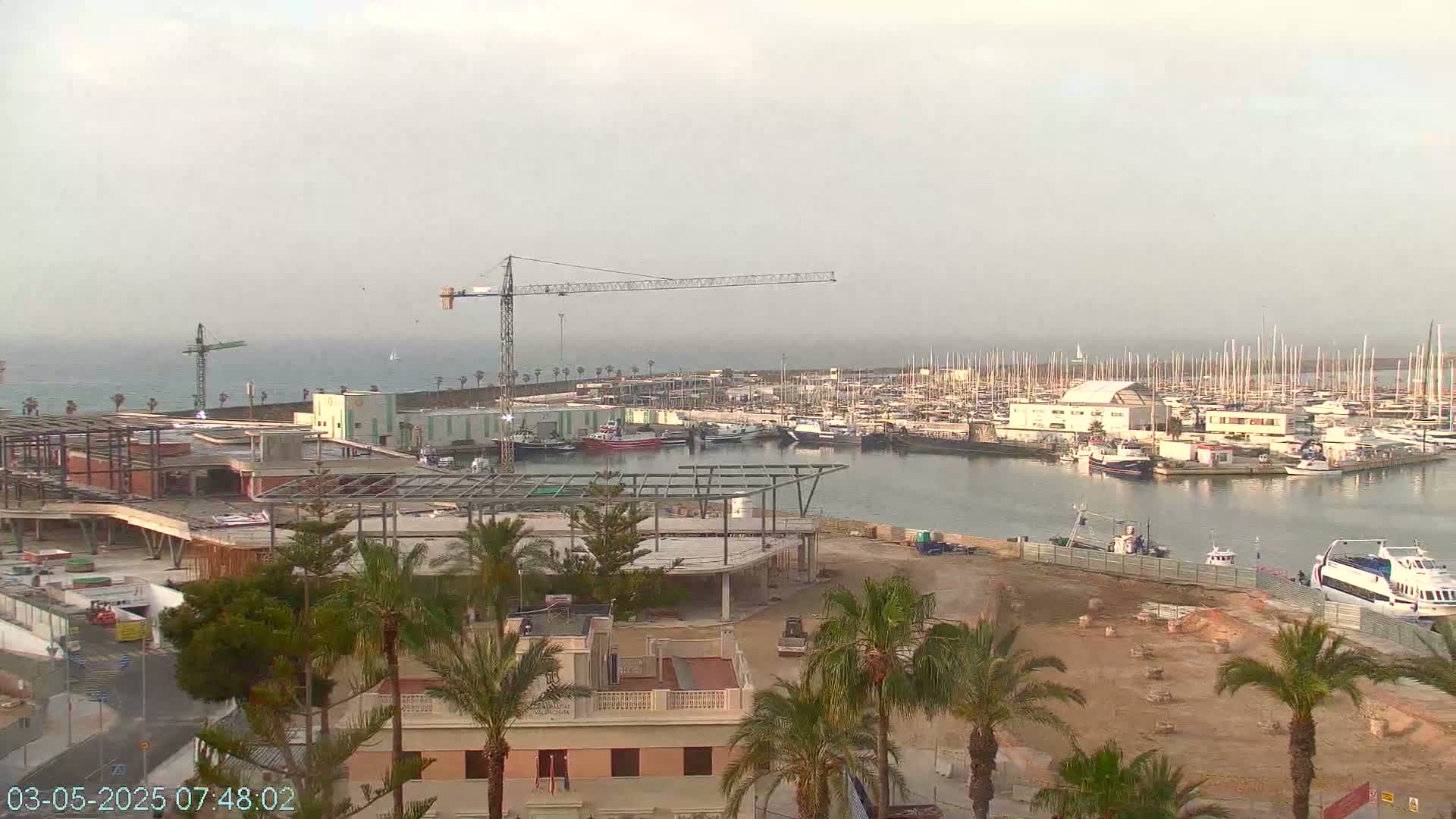 A hazy, overcast day reveals a marina filled with boats, construction underway on a building near palm trees, and construction cranes in the background.
