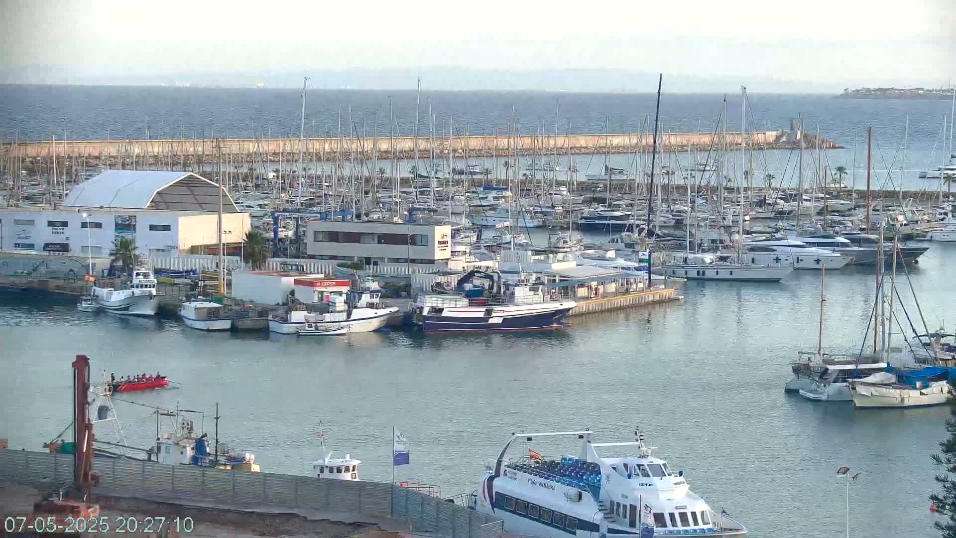 A marina filled with numerous sailboats and motorboats is situated next to calm ocean waters under a mostly clear sky.