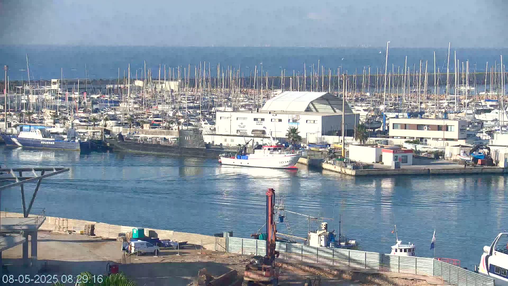A marina filled with numerous sailboats and motorboats under a clear, sunny sky, with a fishing vessel navigating the channel.