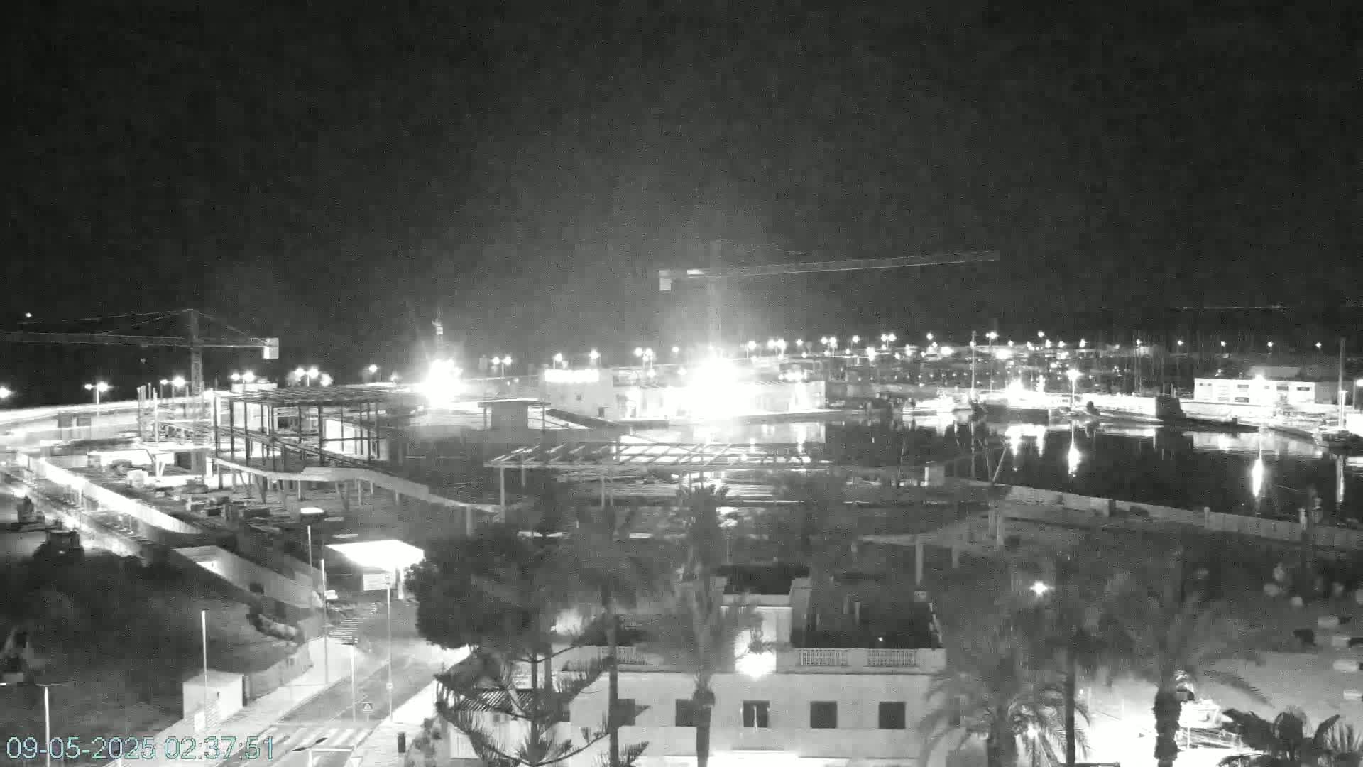 A nighttime, black-and-white view of a construction site next to a marina, illuminated by artificial lights.