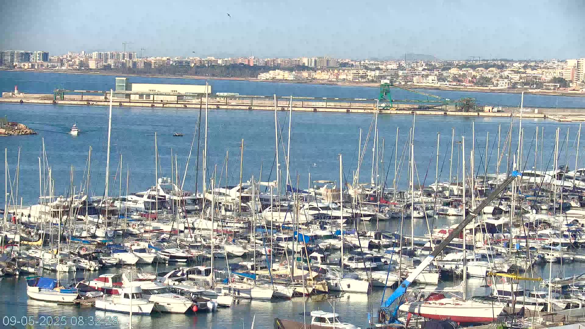A large marina filled with numerous sailboats and motorboats is situated near a city on a calm, sunny day.