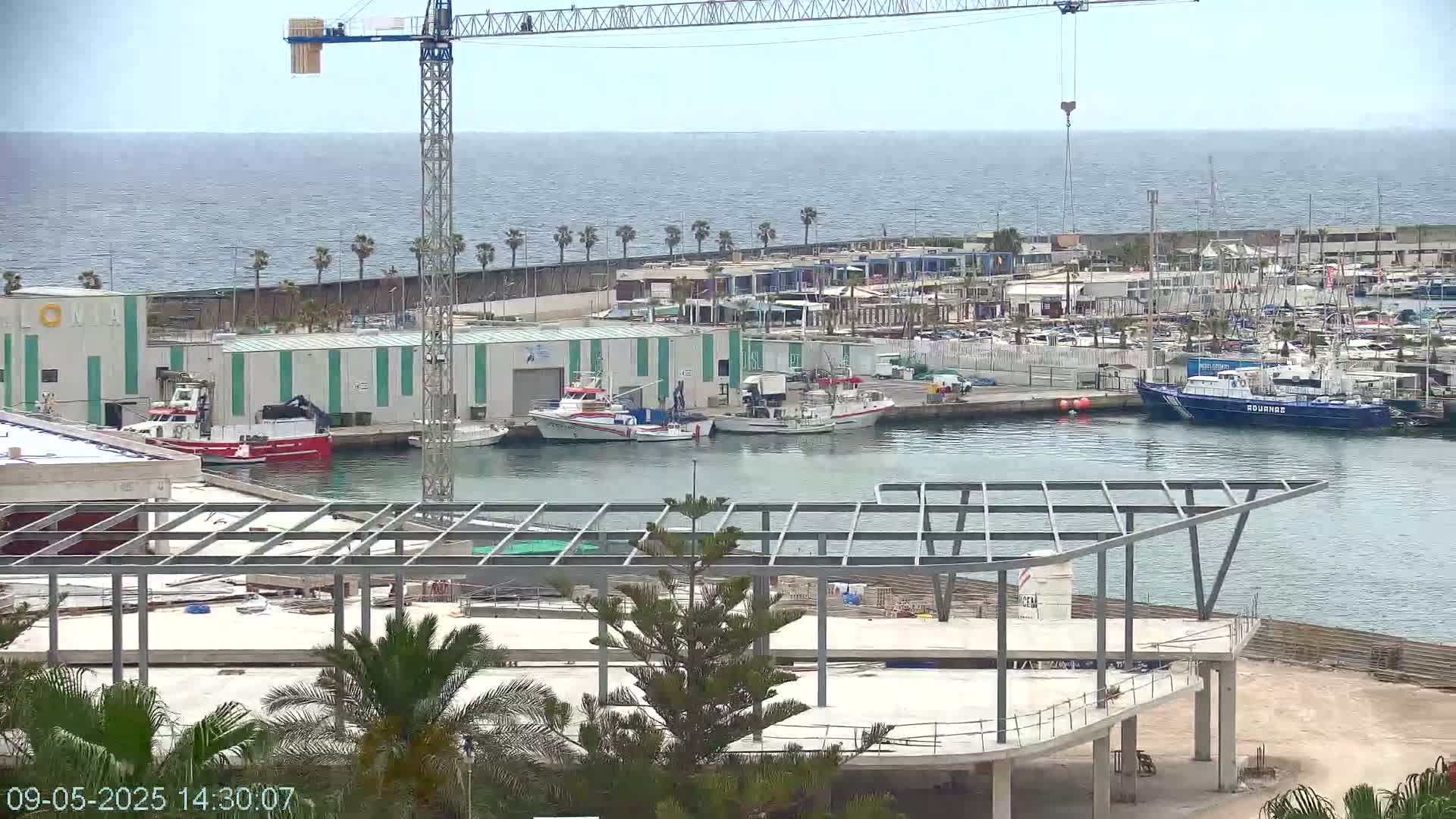 A partially constructed building overlooks a harbor filled with boats under a mostly clear sky, with a crane visible in the background.