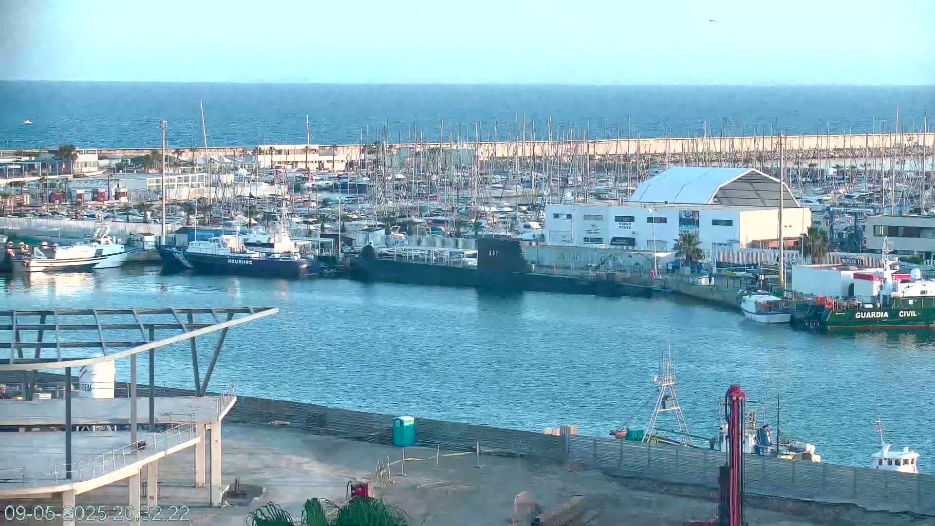 A harbor filled with numerous boats and a submarine, under a clear sky, with calm waters extending to a distant ocean.