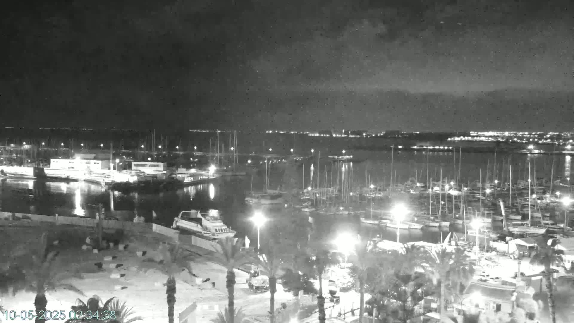 A nighttime, high-angle view of a marina filled with numerous boats and yachts, illuminated by lights, under a dark, cloudy sky.