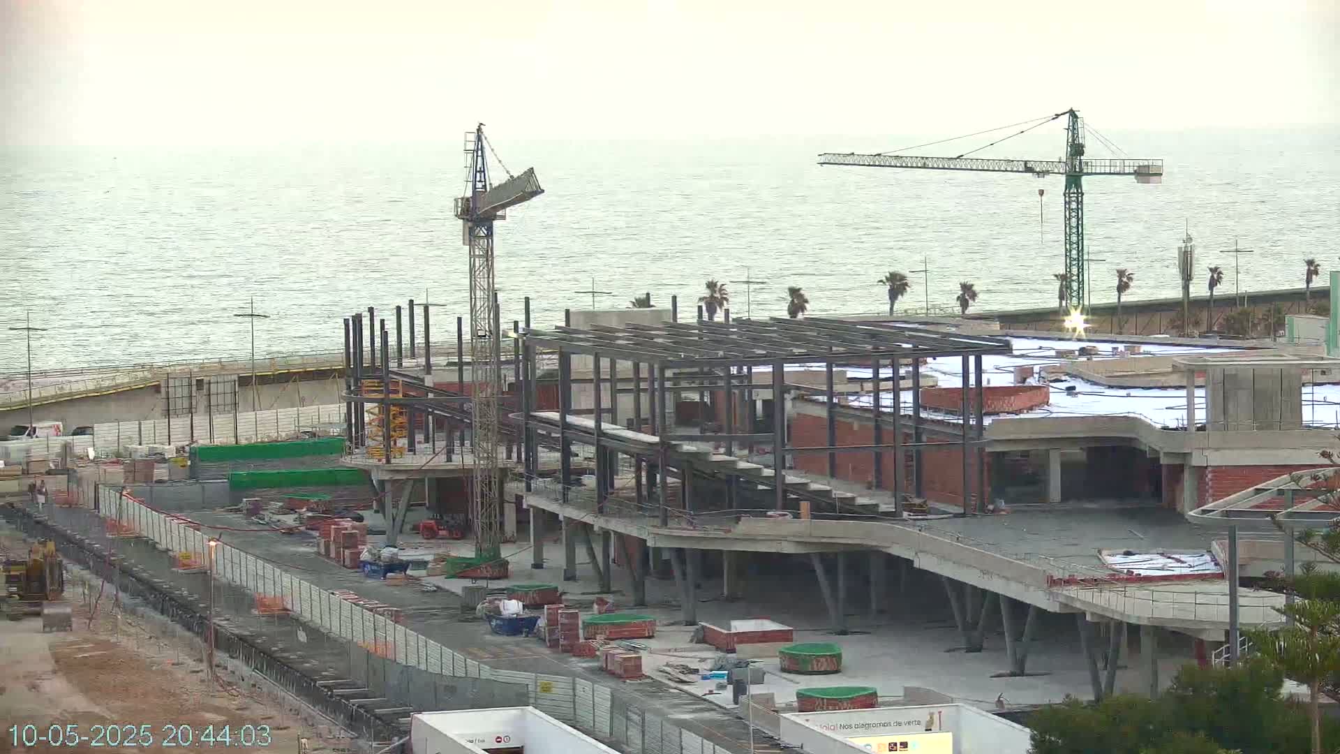 A partially constructed building near a body of water under a cloudy sky, with construction cranes present.