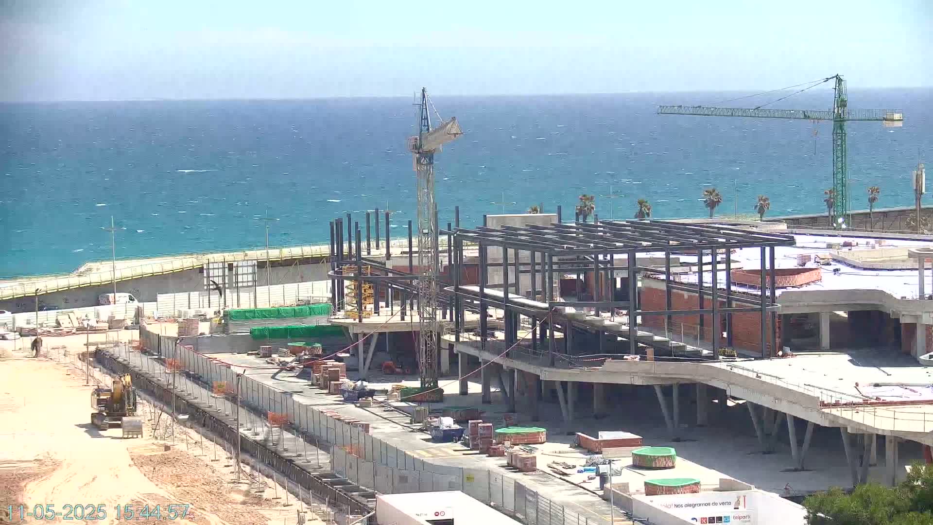 A large building under construction near a wavy ocean on a sunny day, with multiple cranes present.