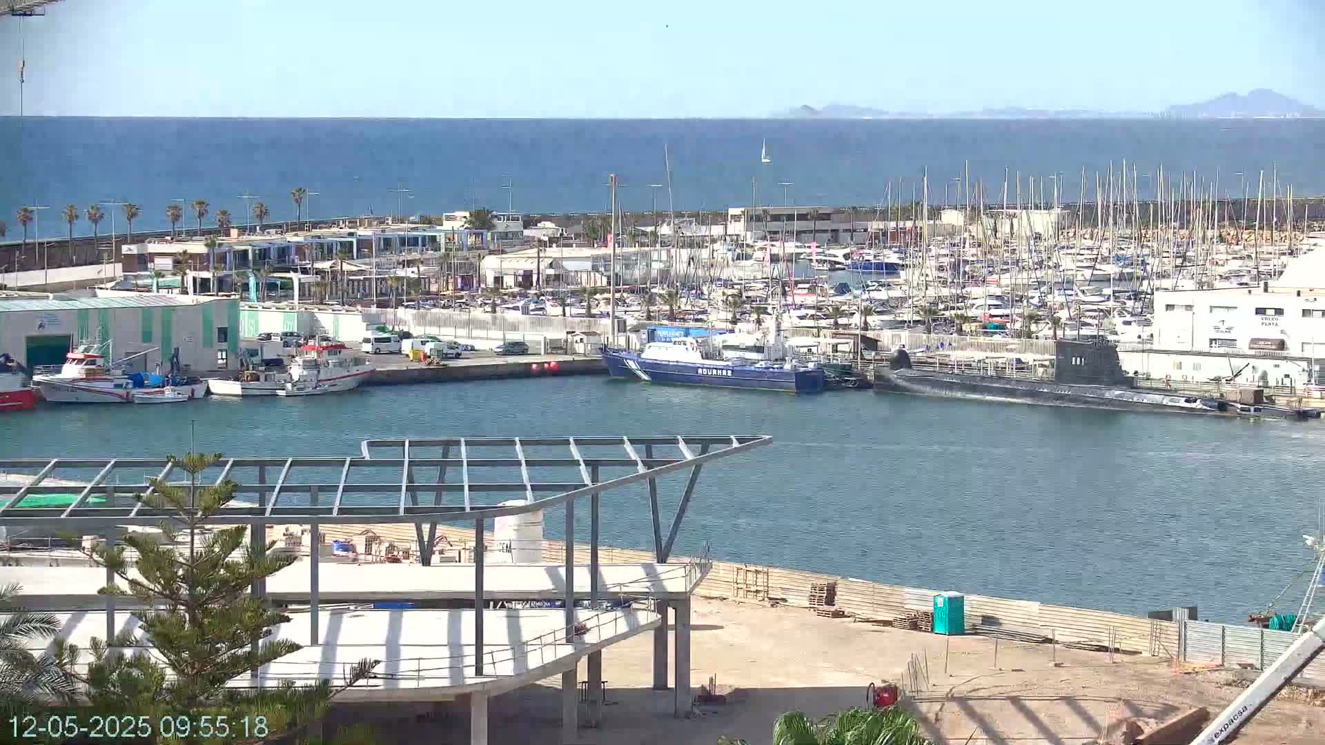 A harbor filled with numerous boats and a docked submarine is seen on a sunny day, with construction underway in the foreground.