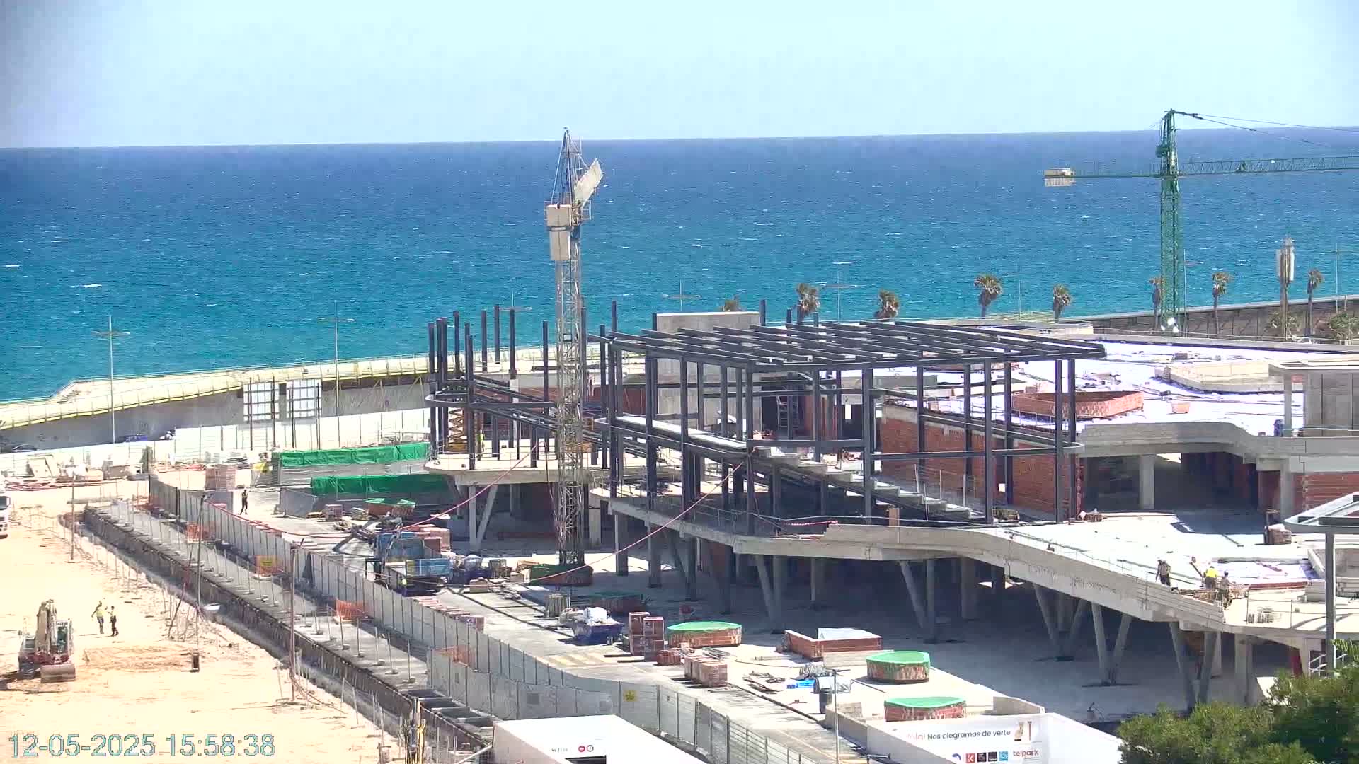 A coastal construction site shows a partially built structure under a clear sky and a calm, blue ocean in the background.