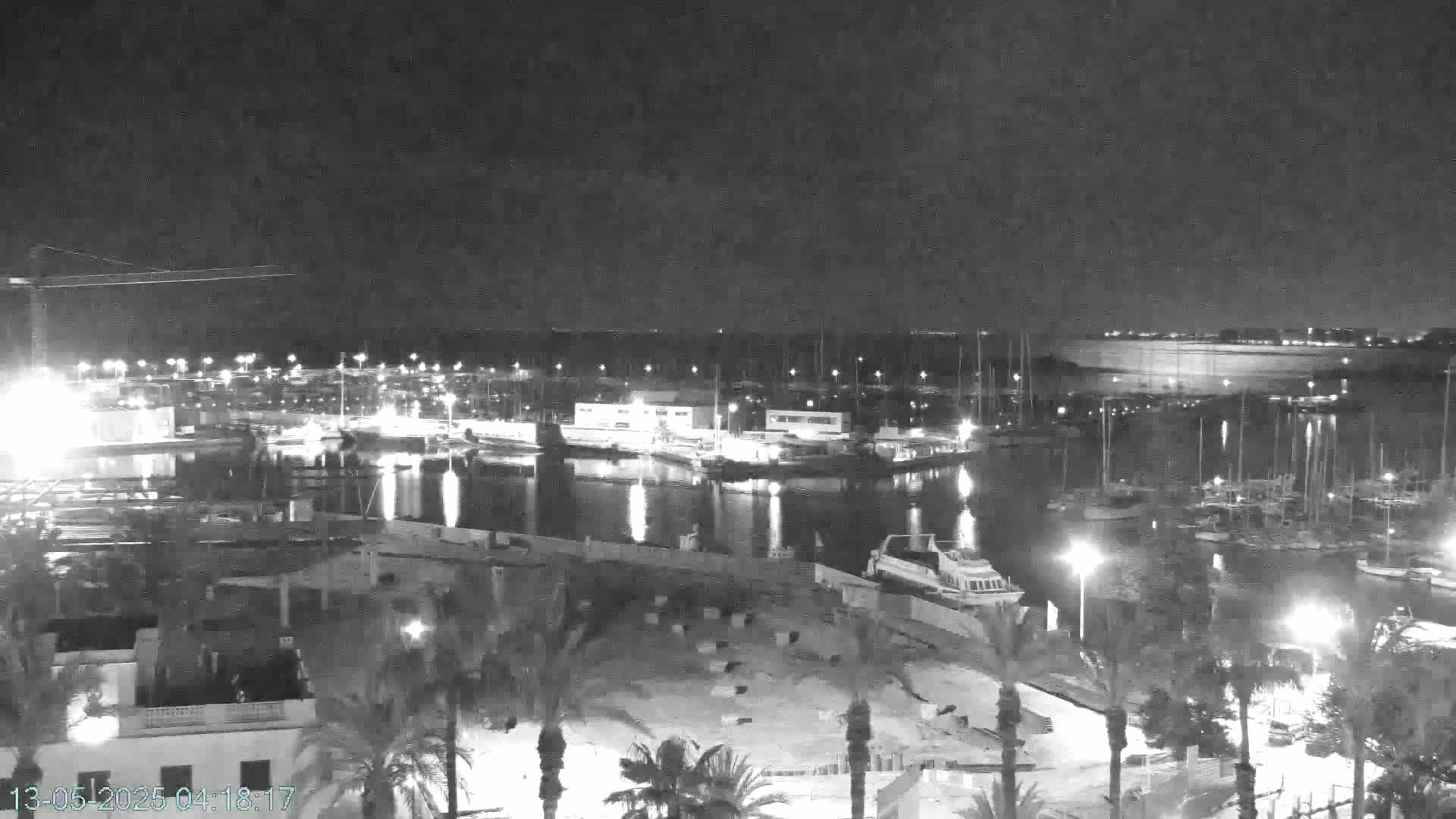 A nighttime view of a marina filled with boats, illuminated by lights, under a dark, possibly cloudy sky.