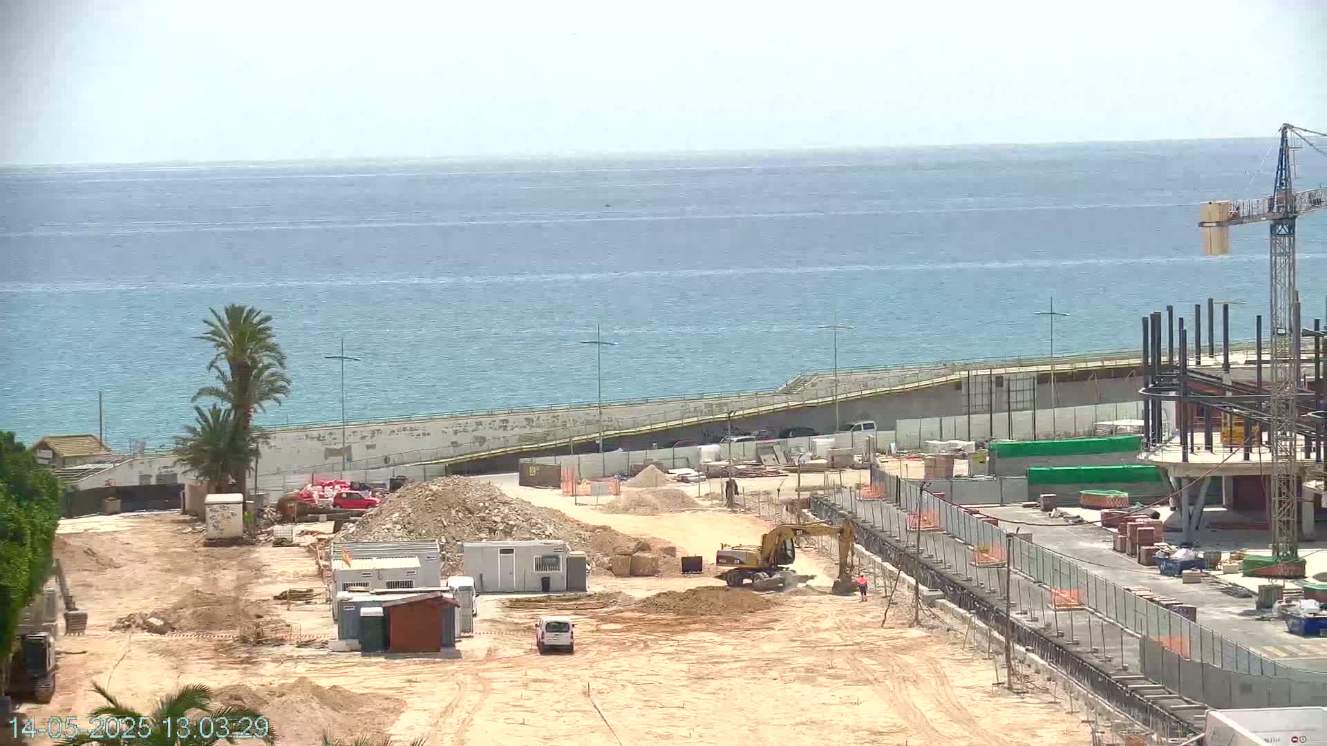 A construction site near a calm ocean is shown under sunny conditions;  heavy machinery and building materials are scattered across the dusty site, with a partially constructed building visible in the background.