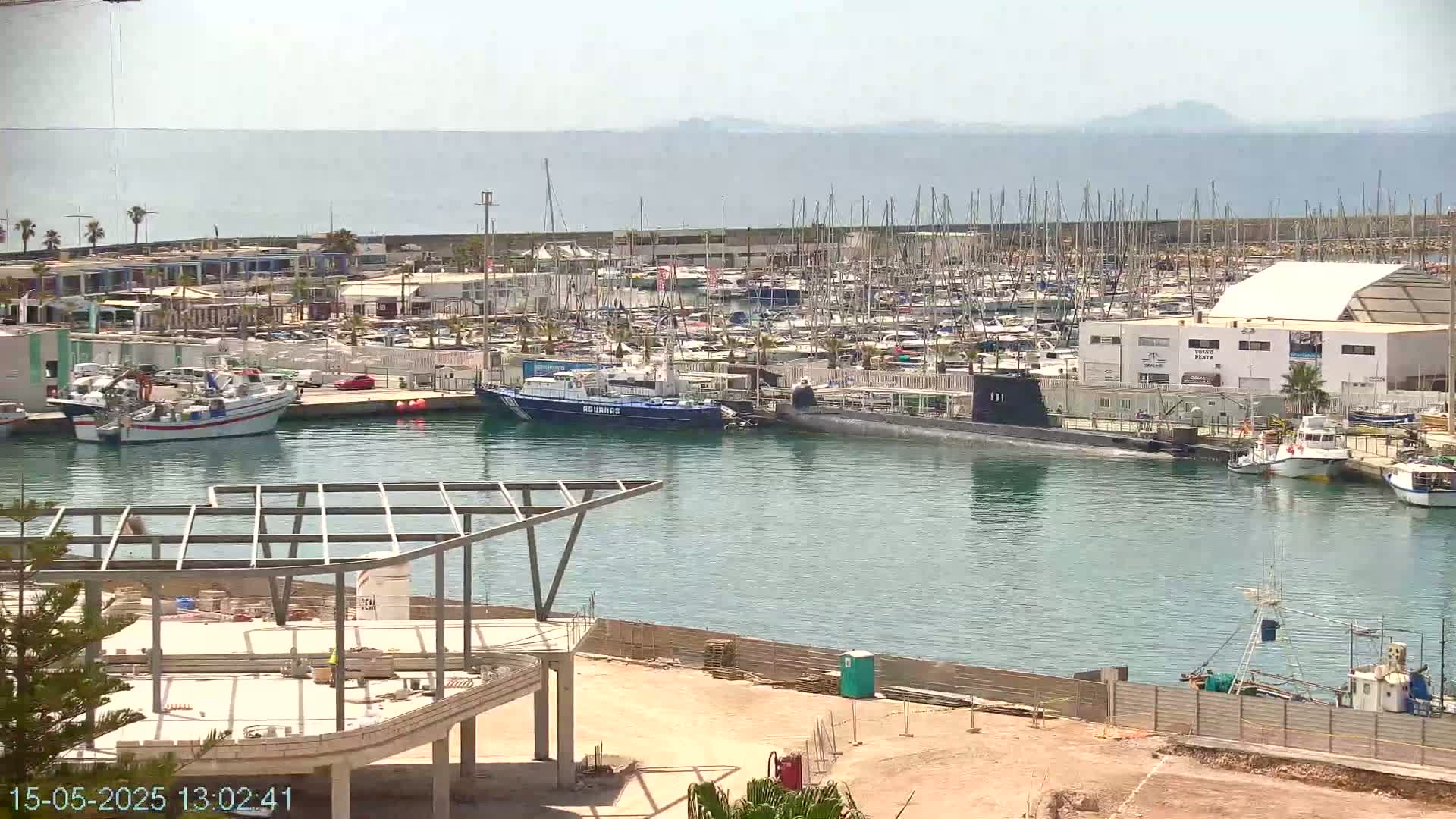 A sunny day at a marina shows numerous boats docked, a submarine, and ongoing construction on the shore.