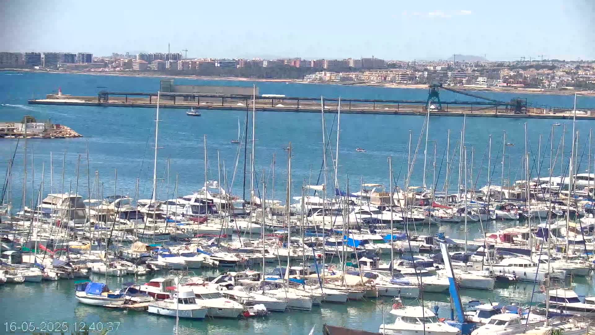 A marina filled with numerous sailboats and motorboats is situated on a calm, sunny day near a coastline with a distant city.