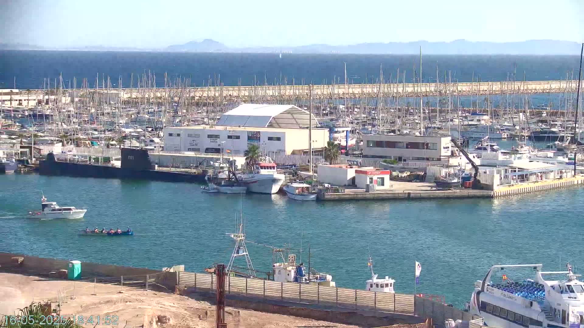 A marina filled with sailboats and motorboats, a submarine, and several fishing vessels is seen on a sunny day with calm water and distant mountains visible on the horizon.