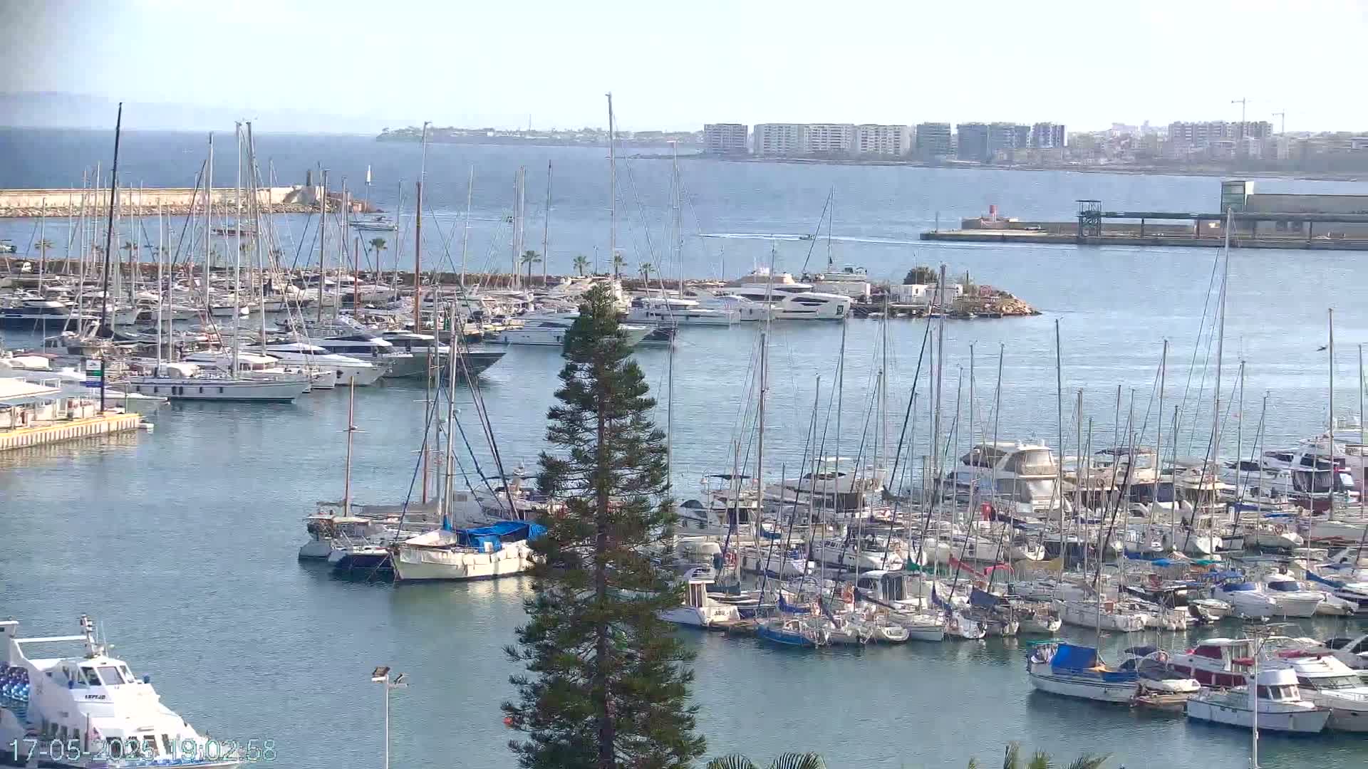 A marina filled with numerous sailboats and motorboats is situated on calm, blue water under a clear sky.
