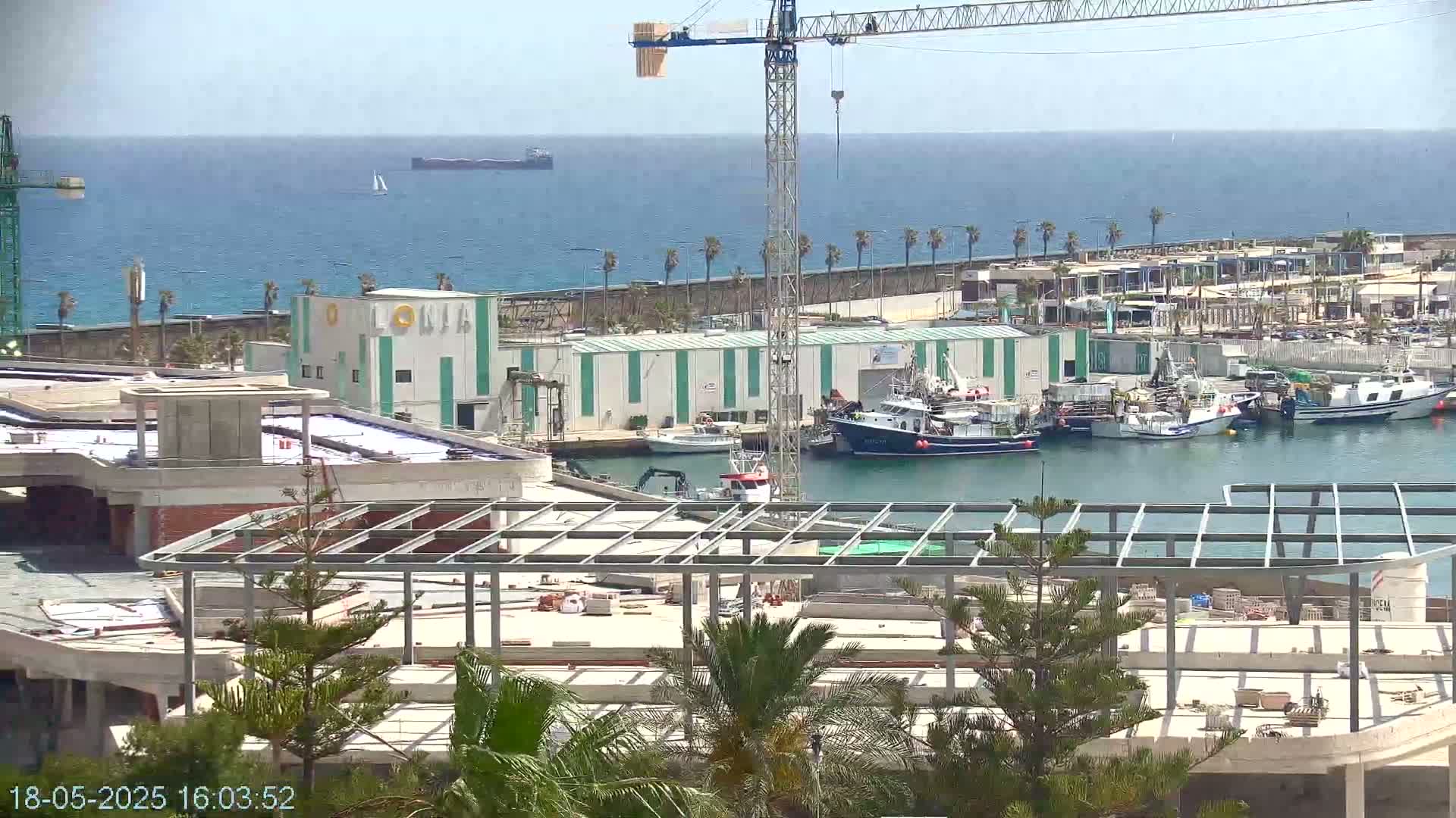 A harbor scene on a sunny day shows a construction site, several fishing boats docked, and a large cargo ship in the distance.