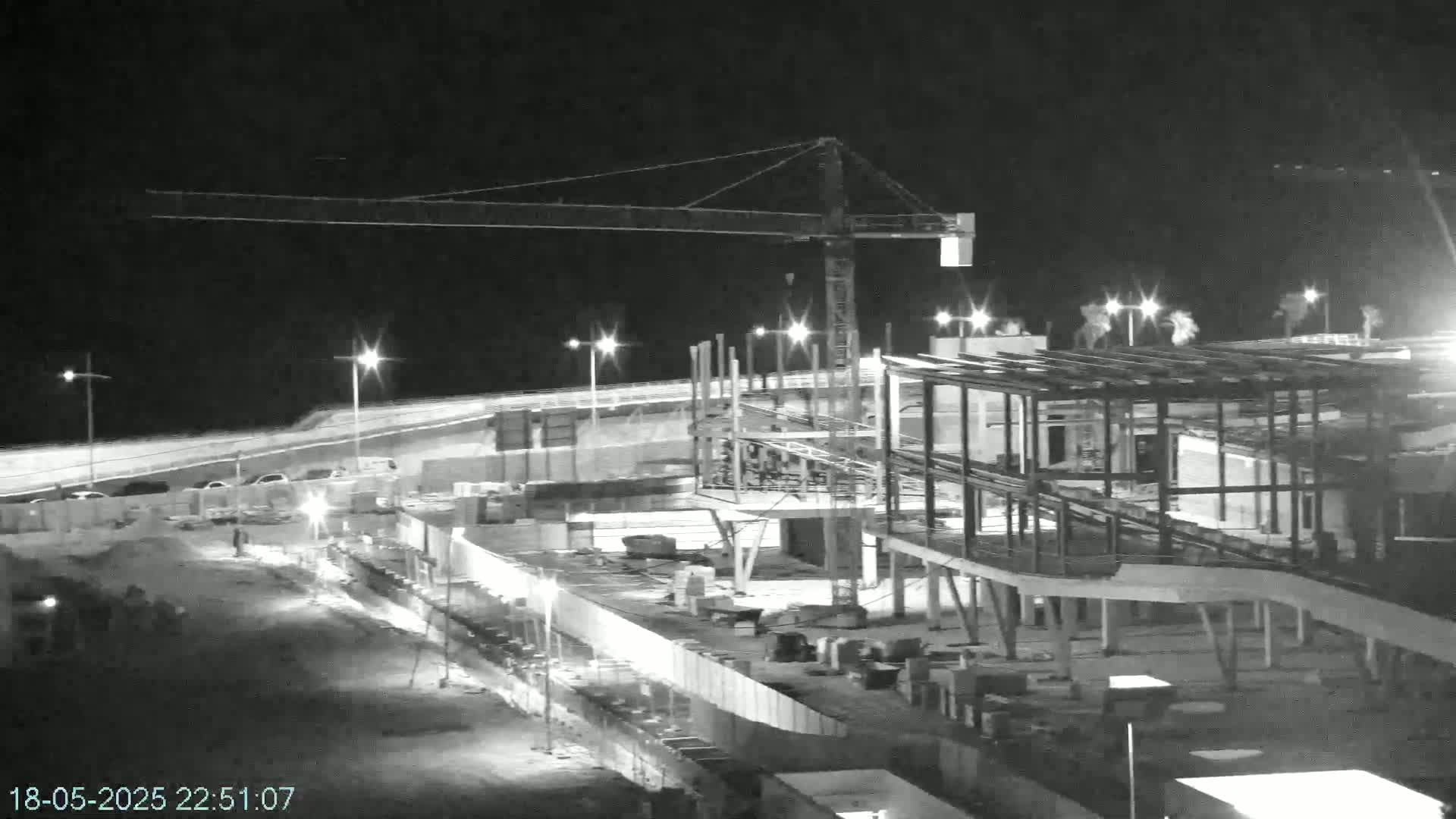 A nighttime, black and white view of a large construction site with a crane, illuminated by artificial lights.