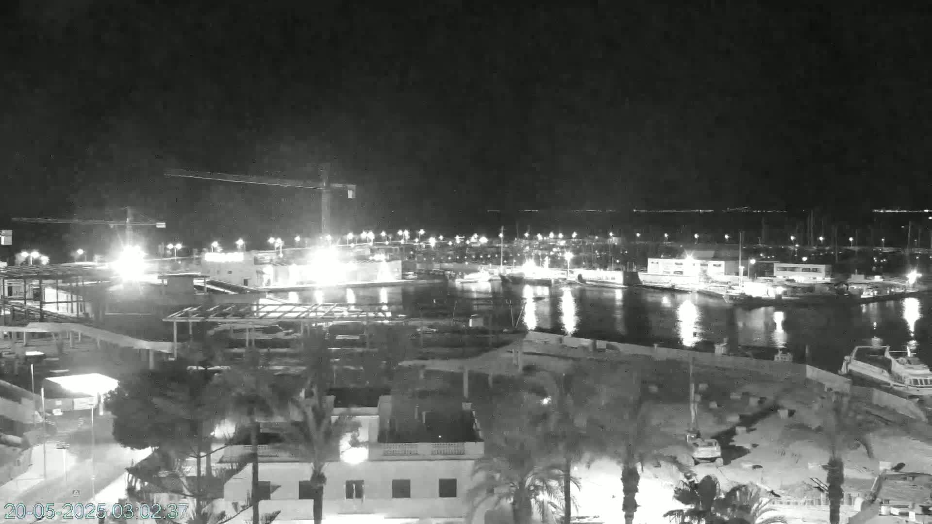 A nighttime, black and white view of a harbor with boats, buildings, and construction underway, under a clear sky.