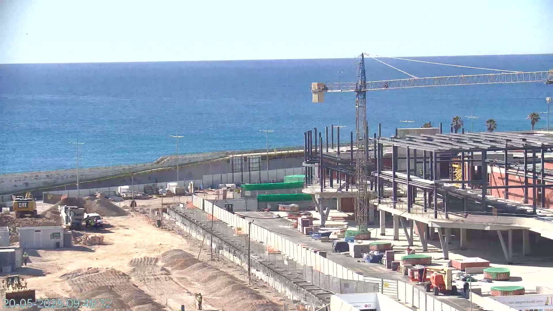 A coastal construction site shows a partially built structure with a crane under a clear, sunny sky overlooking a calm blue ocean.