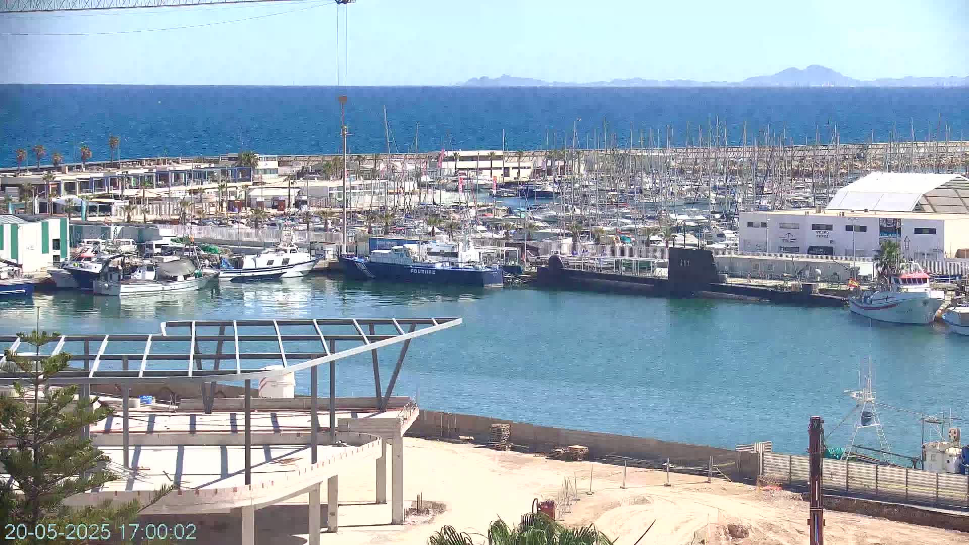 A harbor filled with numerous boats and a docked submarine is seen from an elevated vantage point on a sunny day, with construction underway in the foreground and a calm sea extending to distant mountains in the background.