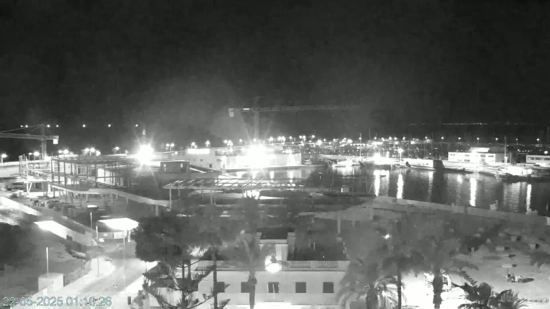 A nighttime, black and white view of a harbor with construction underway near a building, illuminated by lights, under a clear sky.