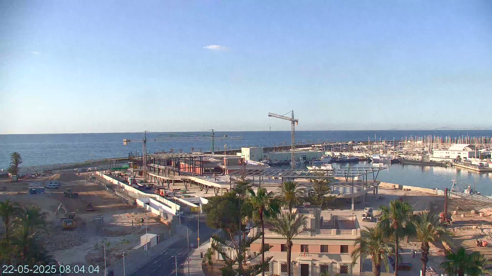 A coastal construction site with several cranes is under a clear, sunny sky, overlooking a marina filled with boats and a calm ocean.