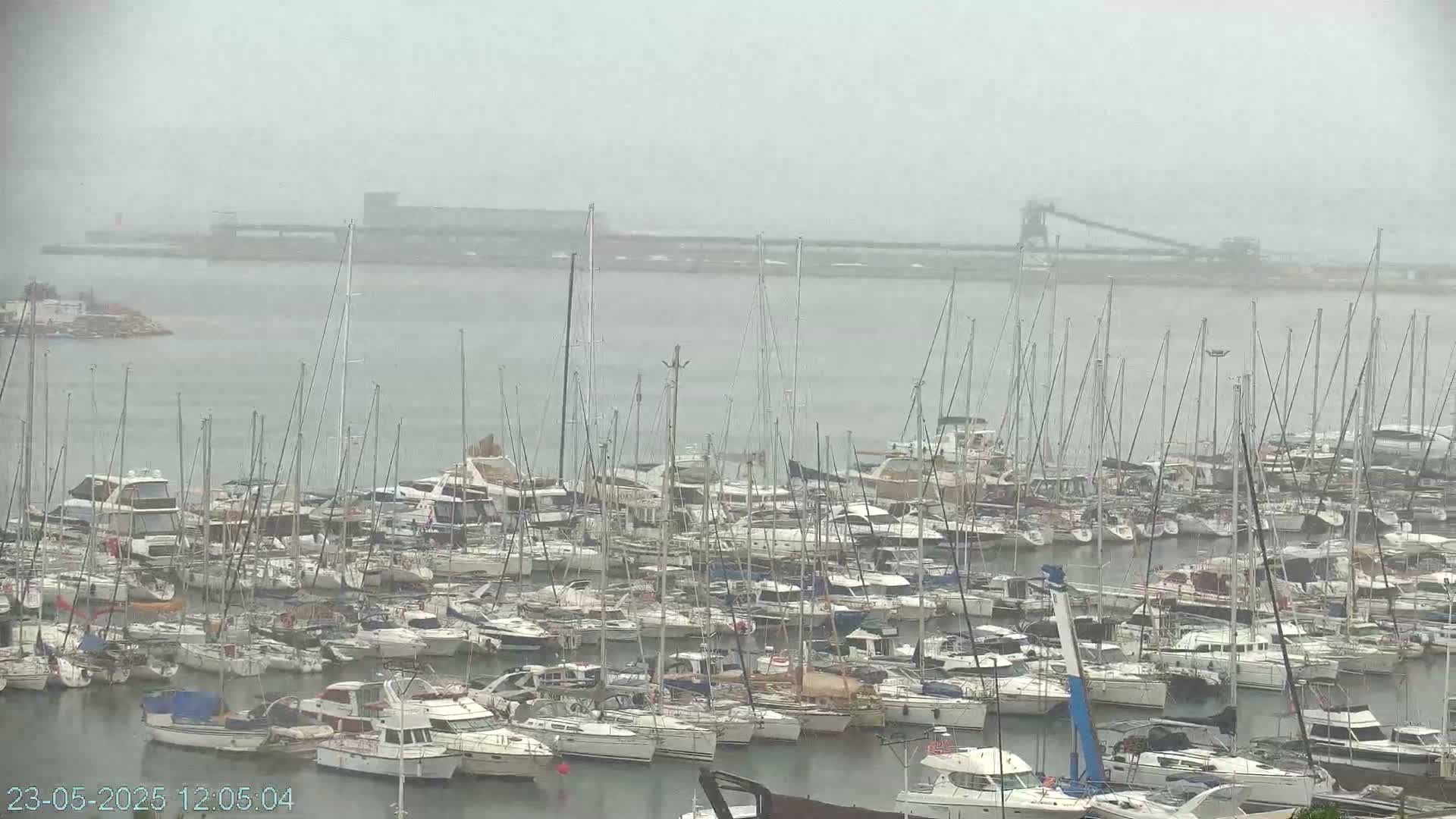 A large marina filled with numerous sailboats and motorboats is seen on an overcast day with misty conditions.