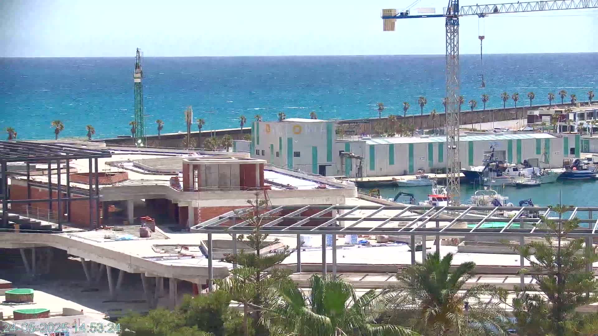 A partially constructed building sits near a harbor filled with boats, under a sunny sky, overlooking a blue ocean with small waves.