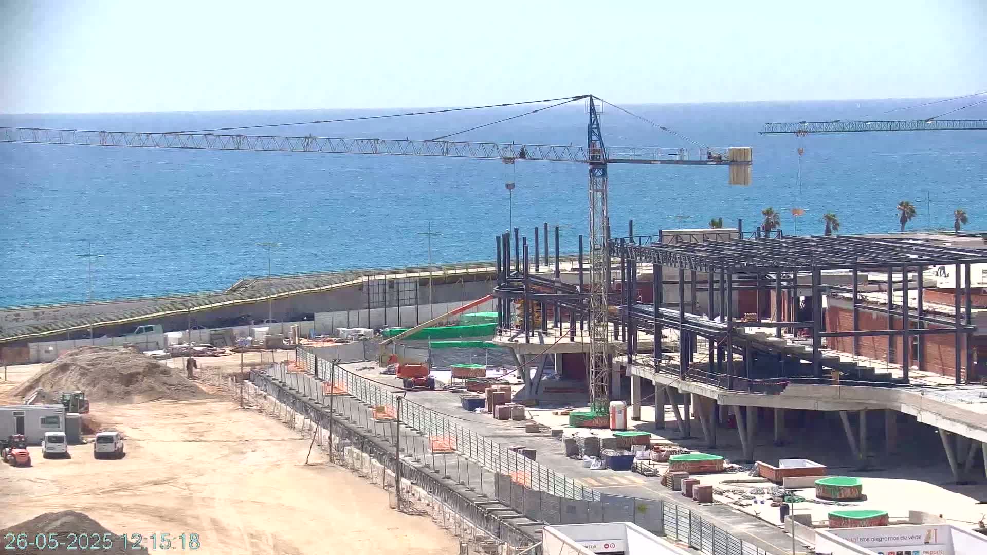 A large building under construction near a body of water on a sunny day, with a crane and various construction vehicles present.