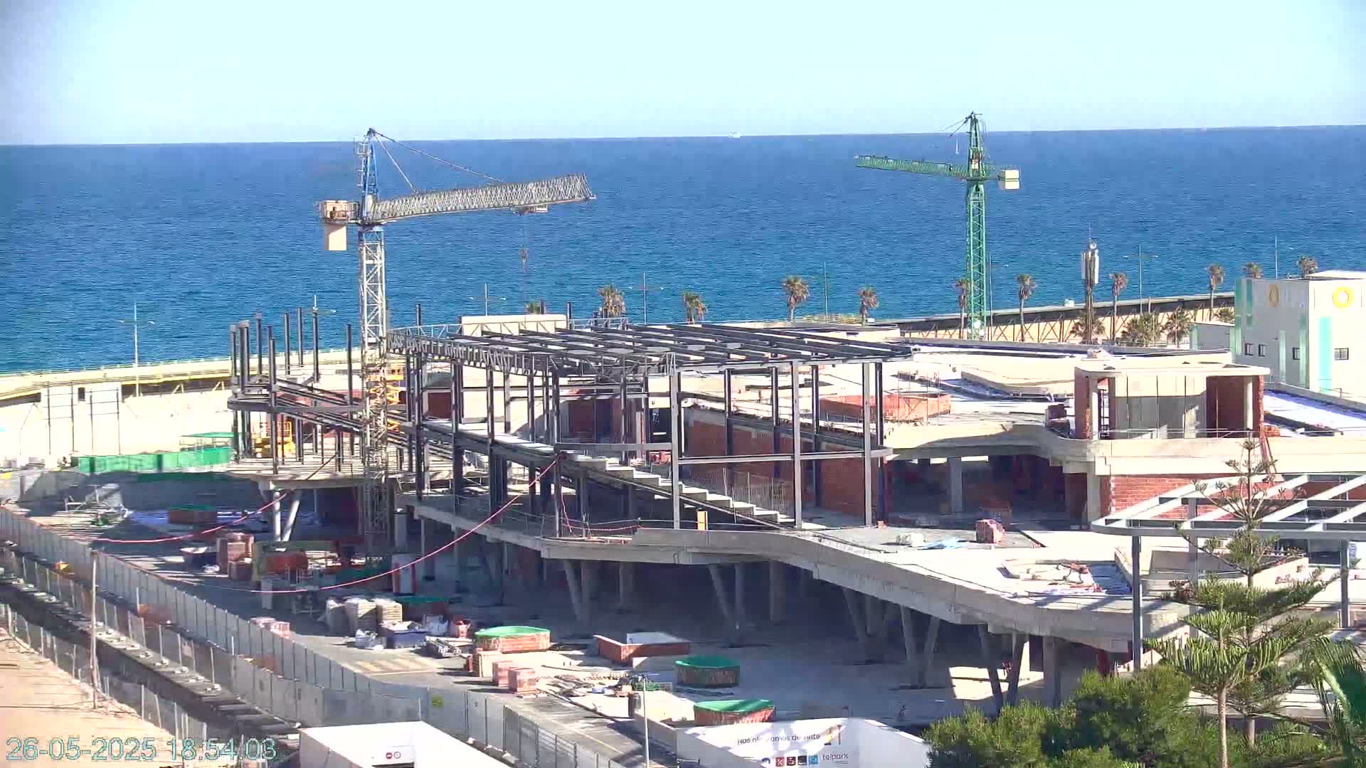 A large building under construction near a body of blue water on a clear day, with two construction cranes visible.