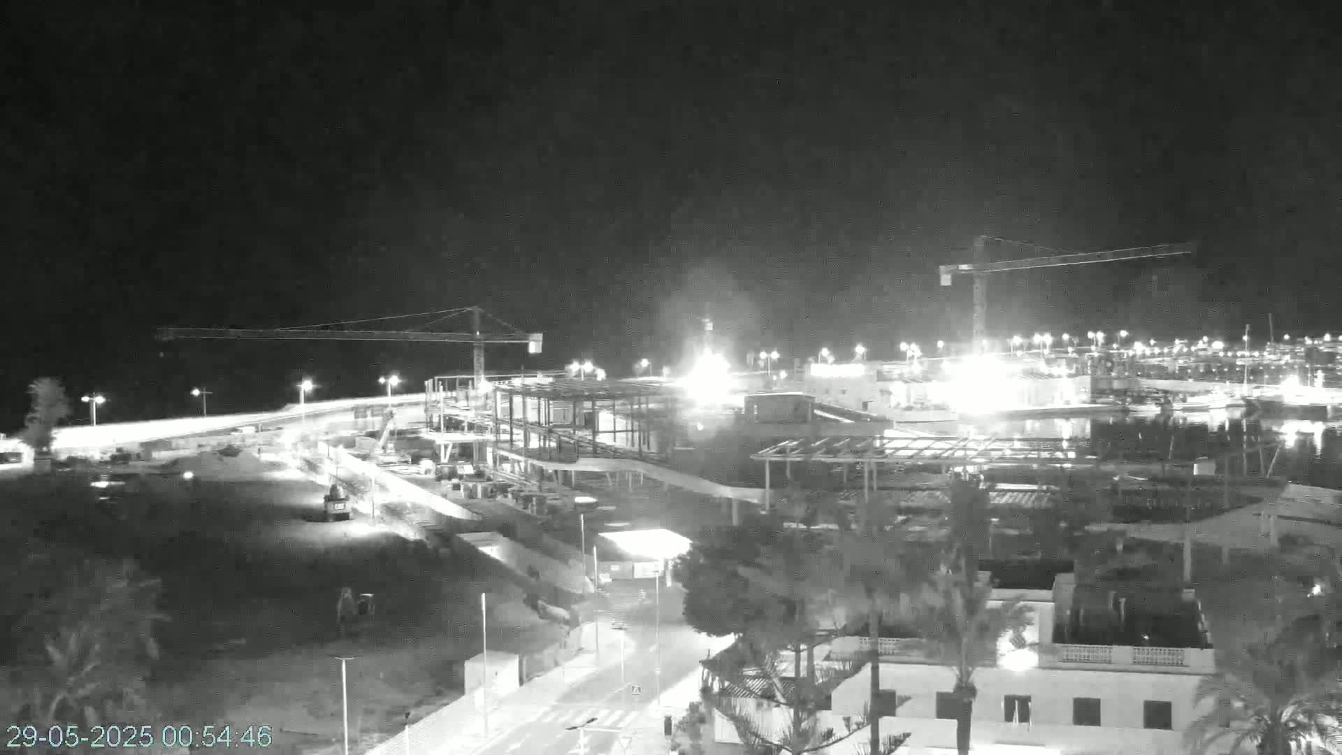 A nighttime, black and white view of a large construction site with cranes, illuminated buildings, and surrounding structures.