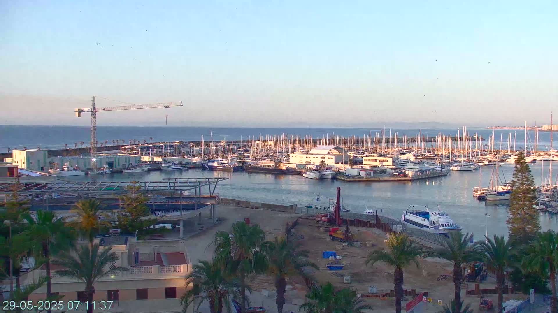 A harbor filled with numerous boats and yachts under a clear, calm sky, with a crane visible on the left and ongoing construction on the lower right.