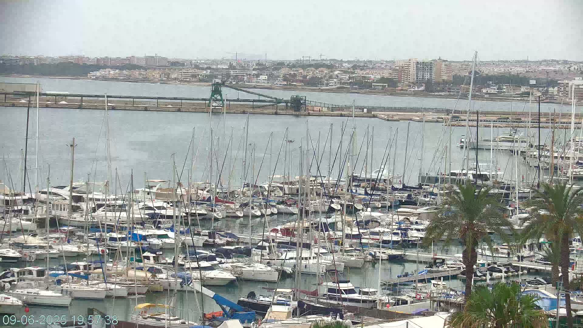 A large marina filled with numerous sailboats and motorboats is situated on calm, gray water under an overcast sky, with a city and industrial structures visible in the background.