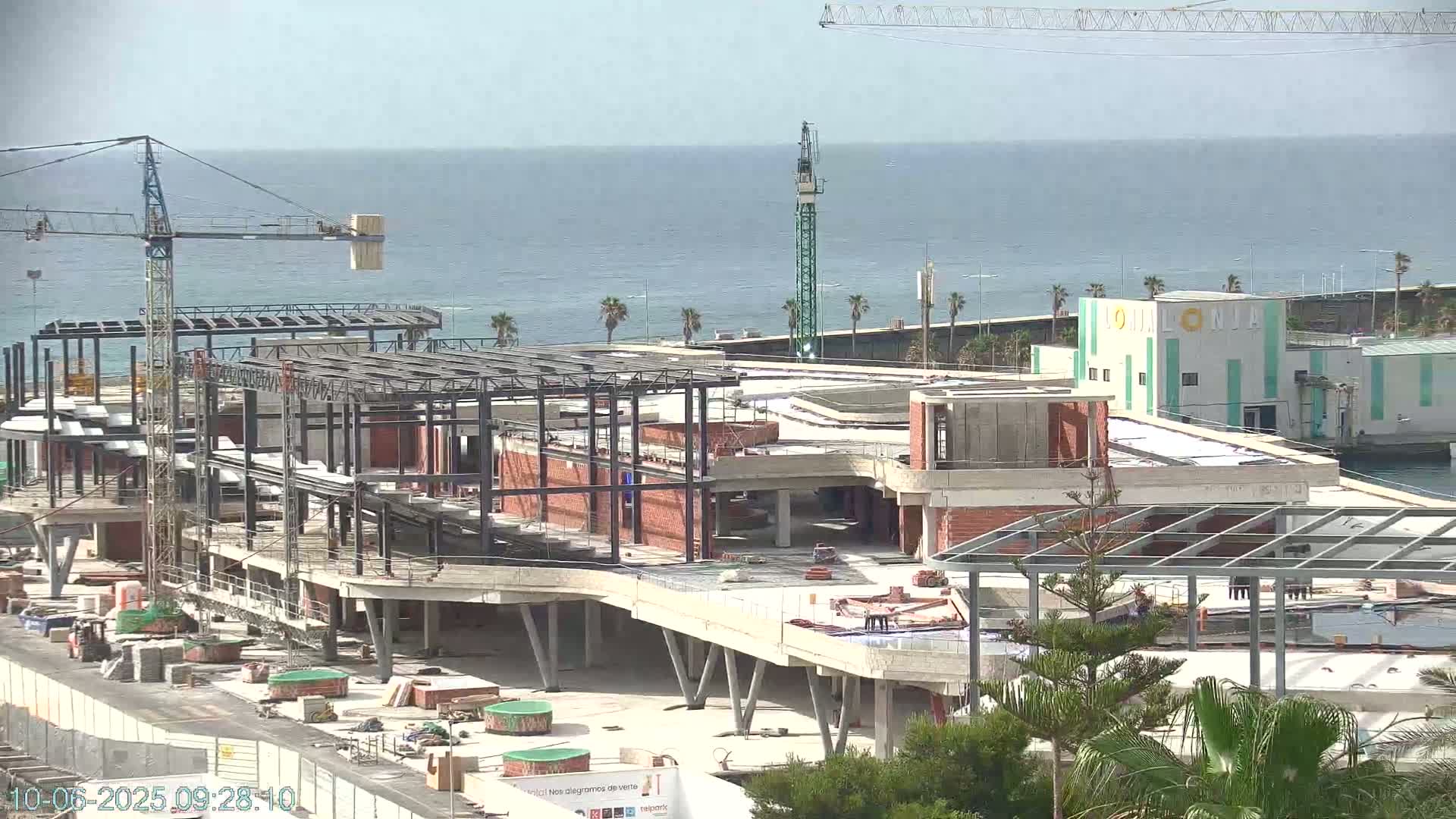 A large building under construction near a body of water on a partly cloudy day, with several cranes visible on the site.