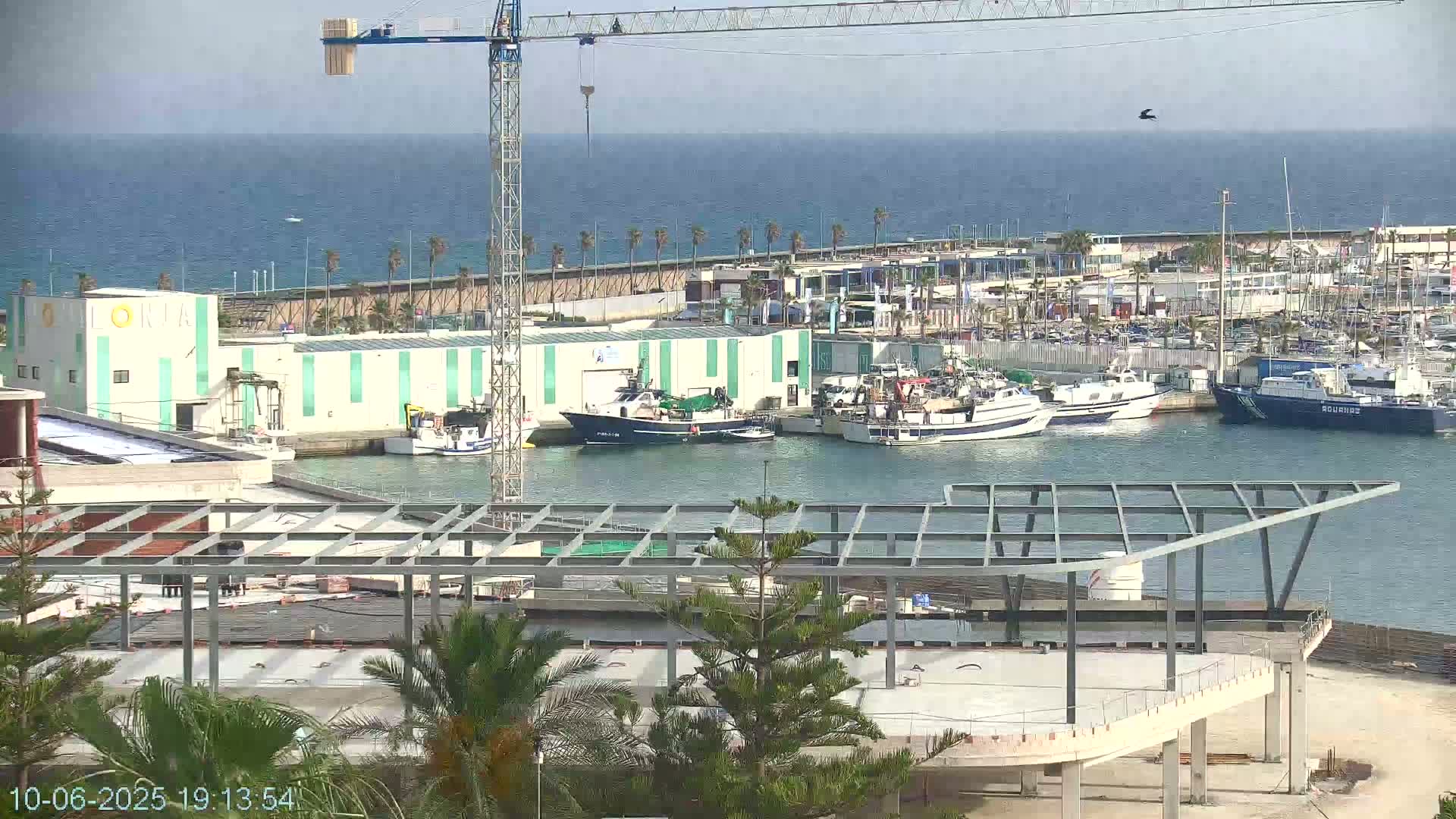 A harbor filled with fishing boats is seen under a clear sky, with a crane and a partially constructed structure in the foreground.