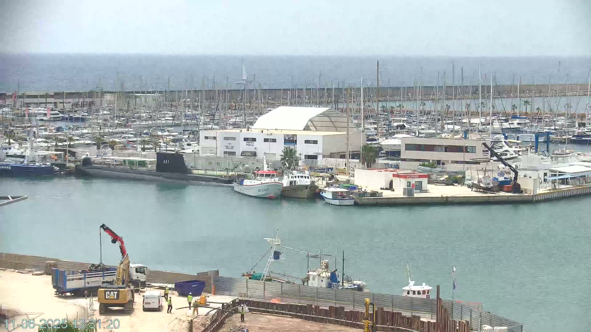 A marina filled with numerous boats and yachts, including a submarine, under a mostly clear sky, with construction work underway on the lower left.