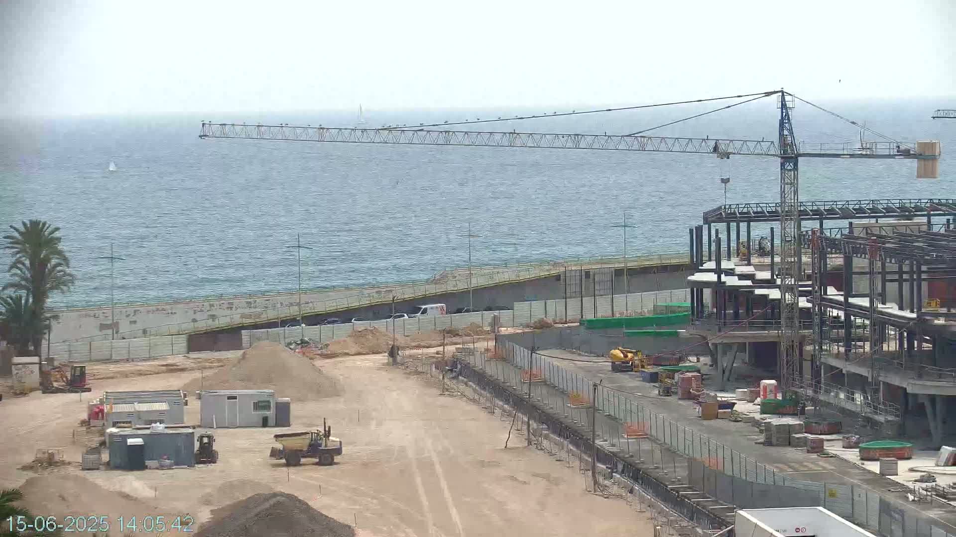 A large construction site near the ocean under a mostly cloudy sky shows a crane, building framework, and various pieces of construction equipment.