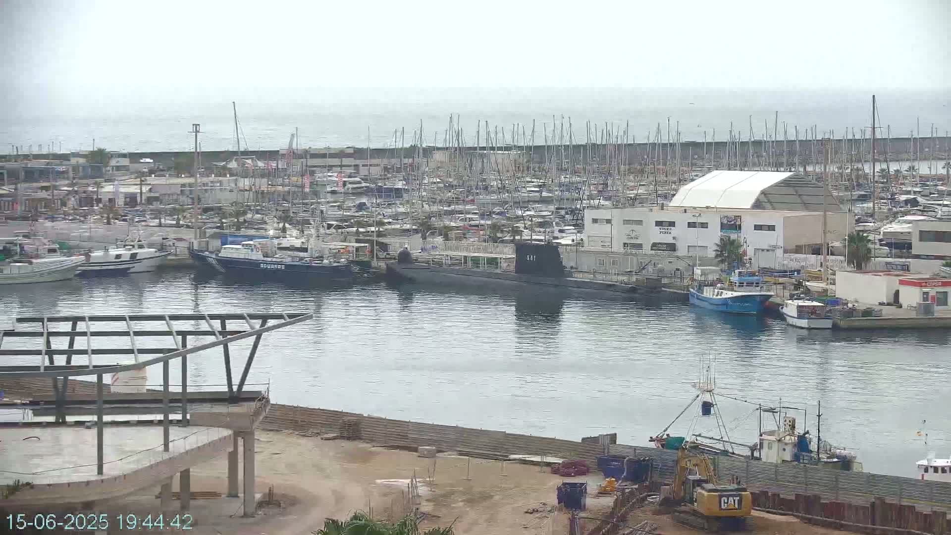 A dark-colored submarine is moored in a harbor filled with numerous boats and yachts under an overcast sky.