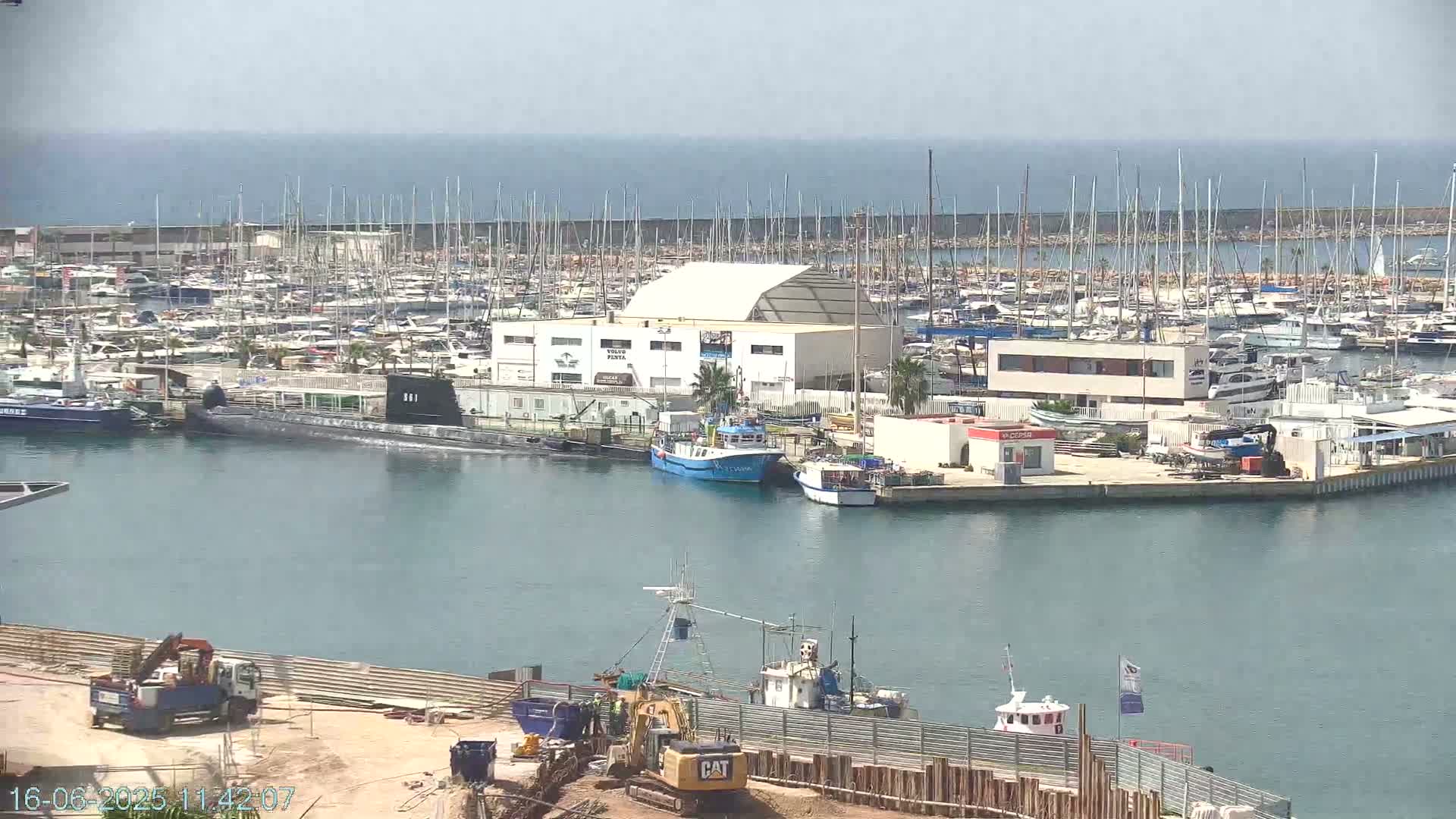 A harbor filled with numerous boats and yachts under a clear sky, with a submarine and construction work visible in the foreground.
